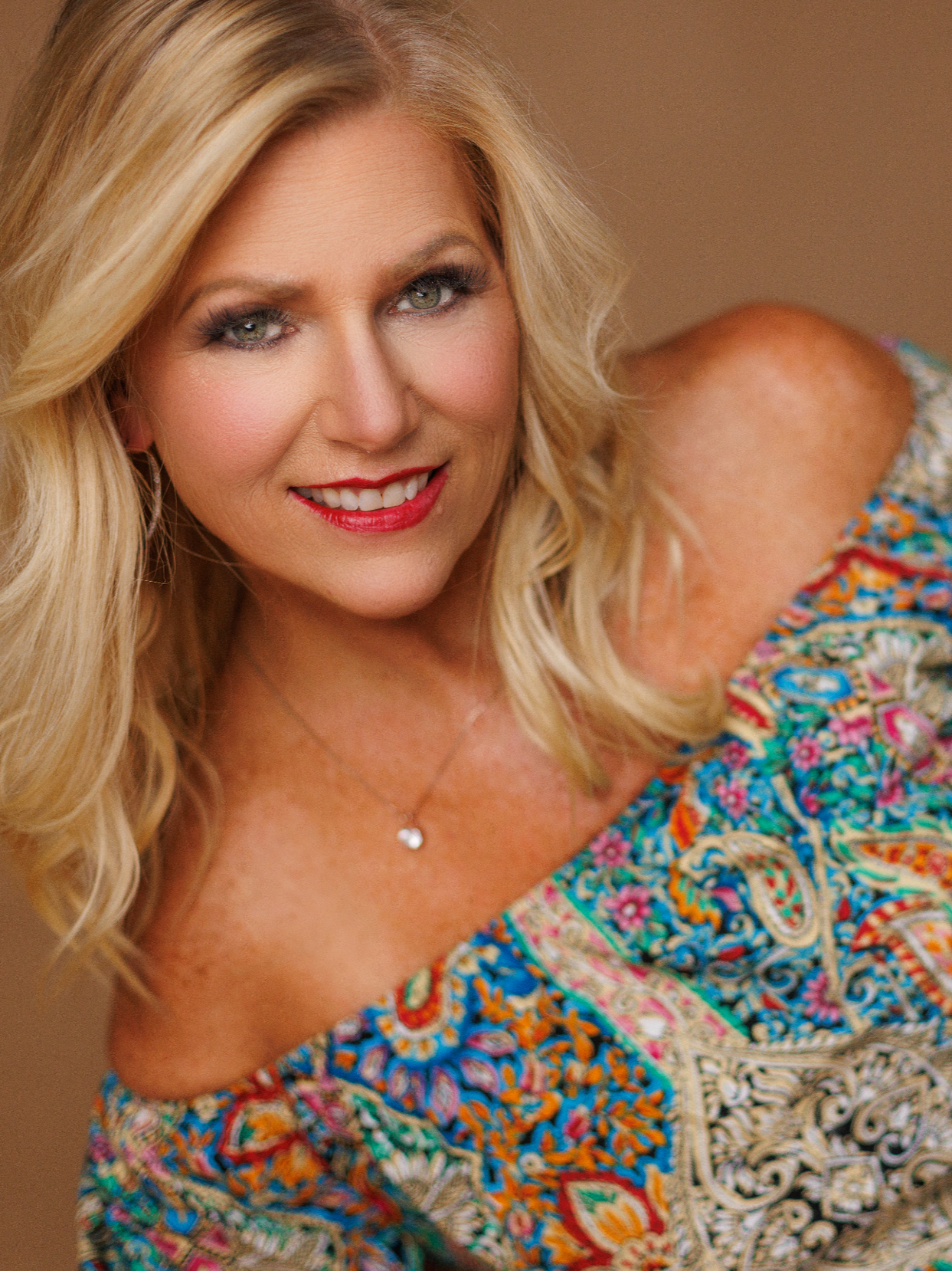 A middle-aged woman with blonde hair smiling, wearing a colorful patterned off-shoulder top and dark jeans, lying on her side against a plain brown background.