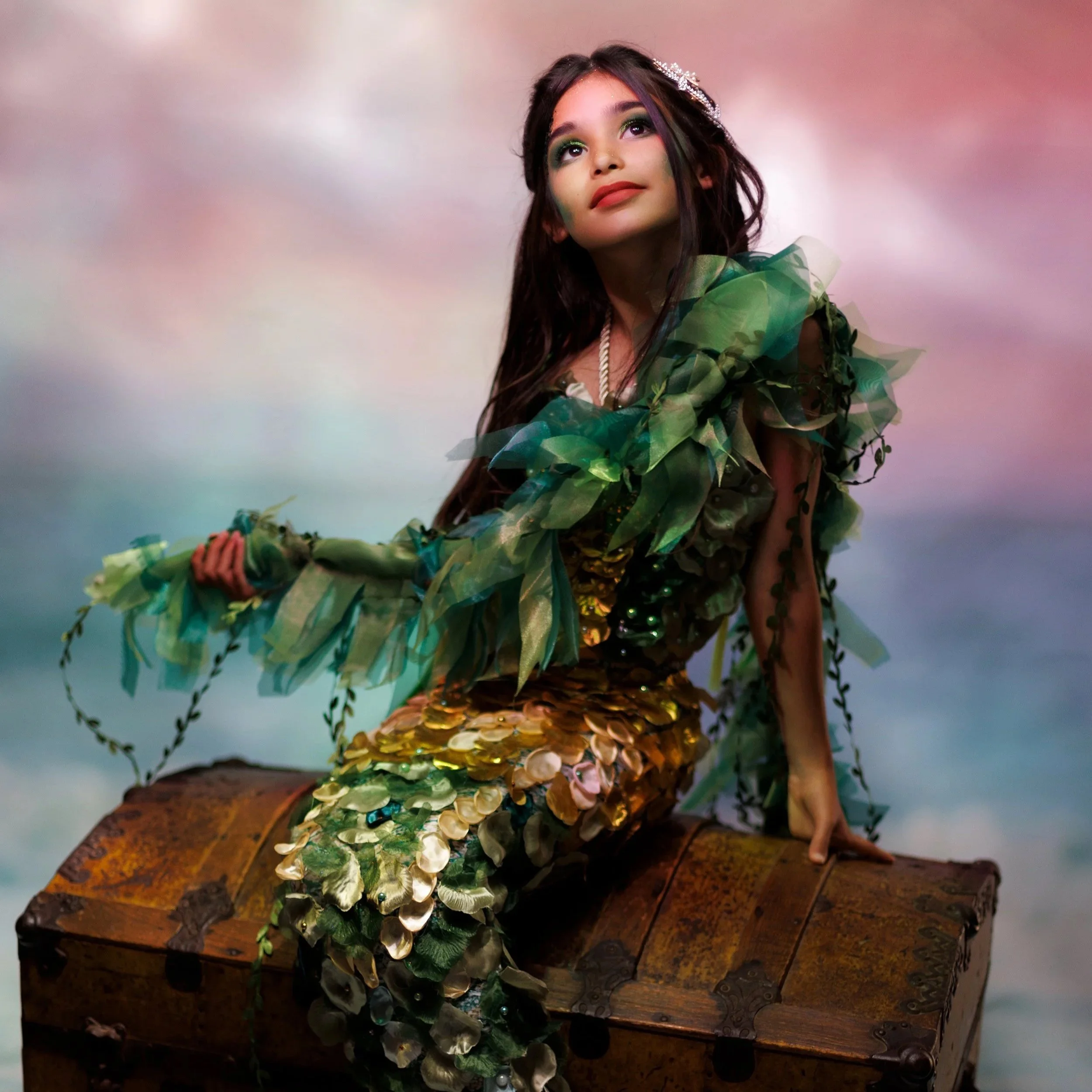 Young girl dressed as a mermaid, sitting on an old wooden chest with a cloudy, colorful sky in the background.
