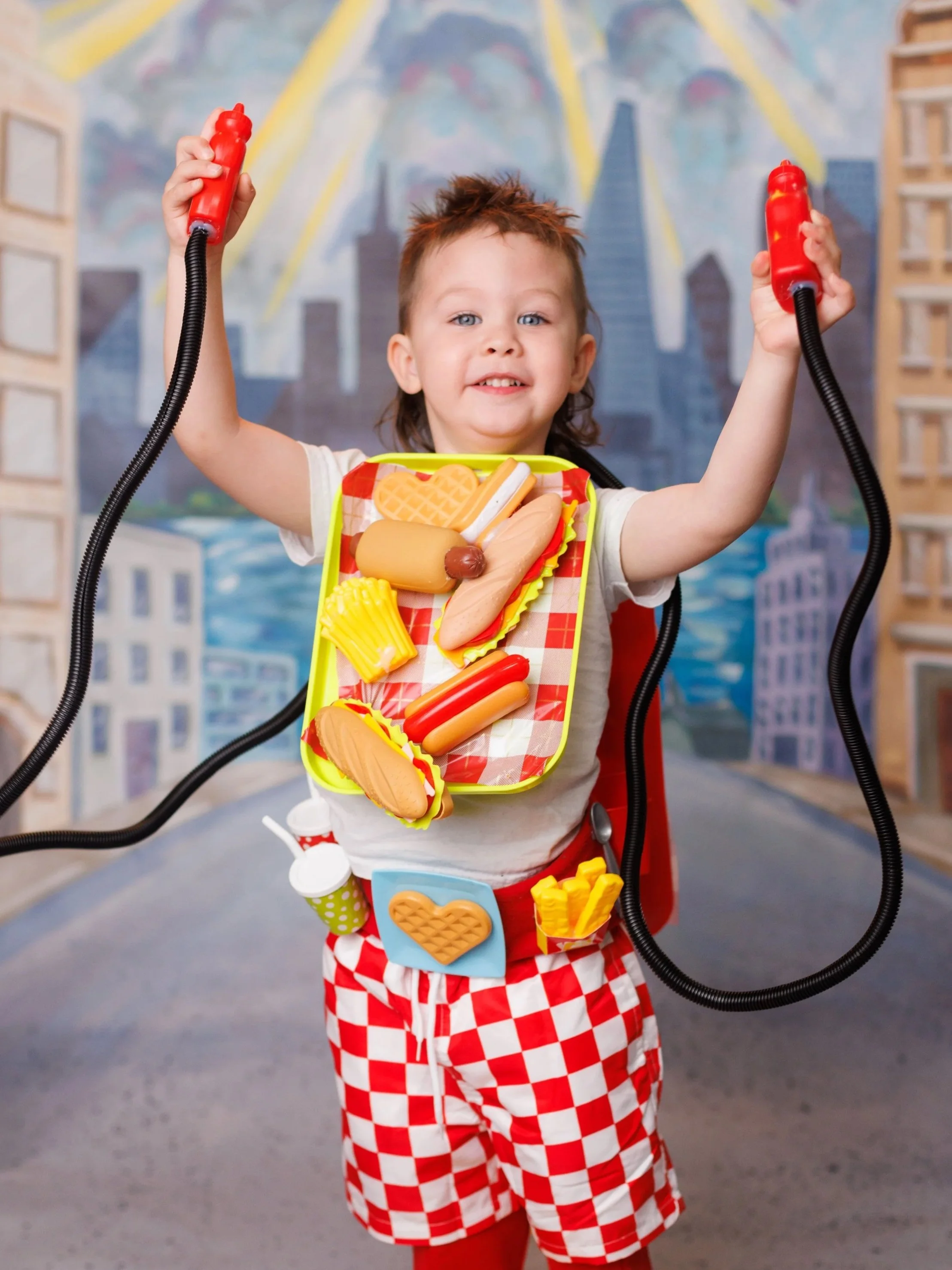 Child dressed as a hot dog vendor holding toy condiments and play food in a city street setting with a cityscape backdrop.
