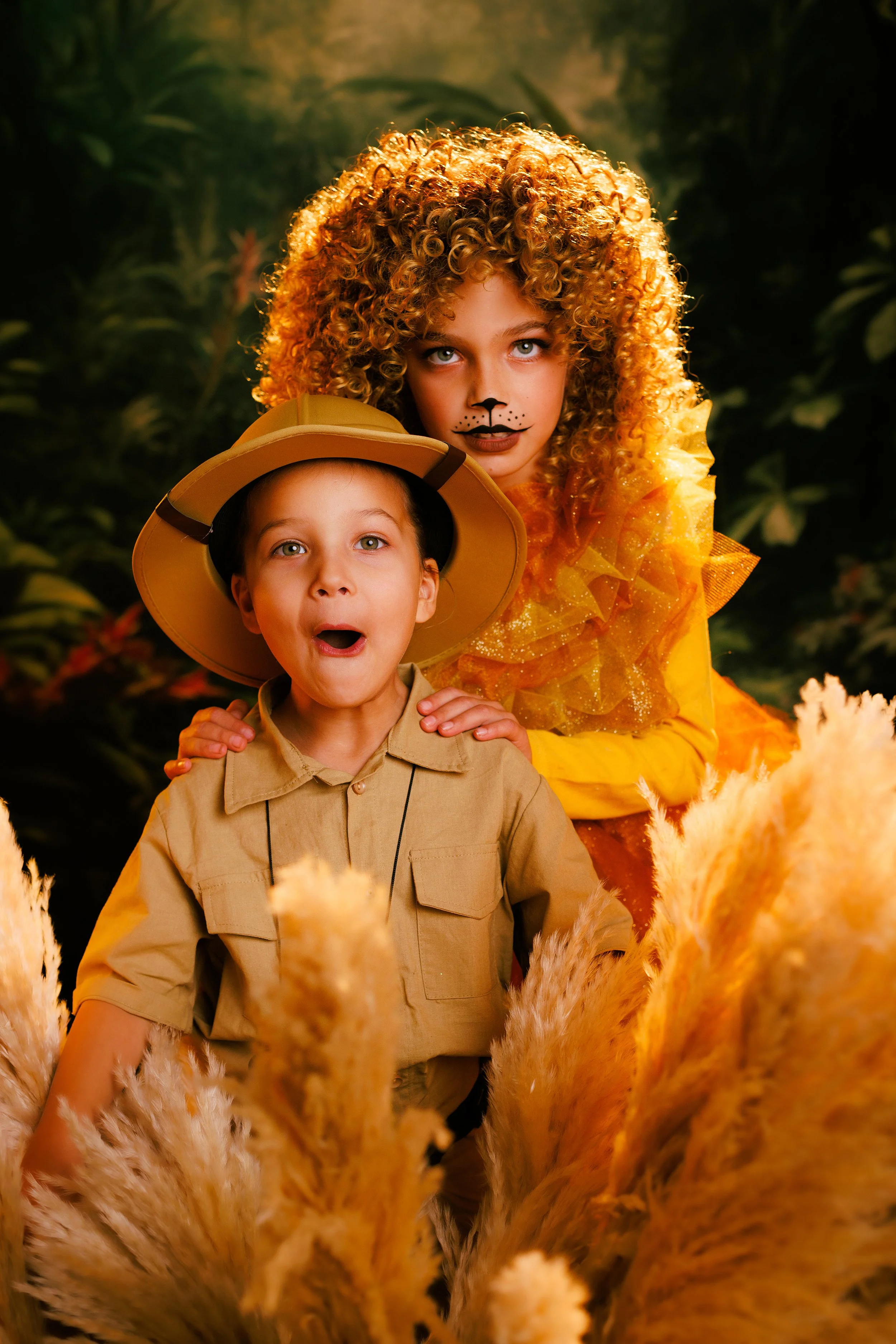 Two children dressed as jungle explorer and lion, surrounded by tall pampas grass, in a lush jungle setting.