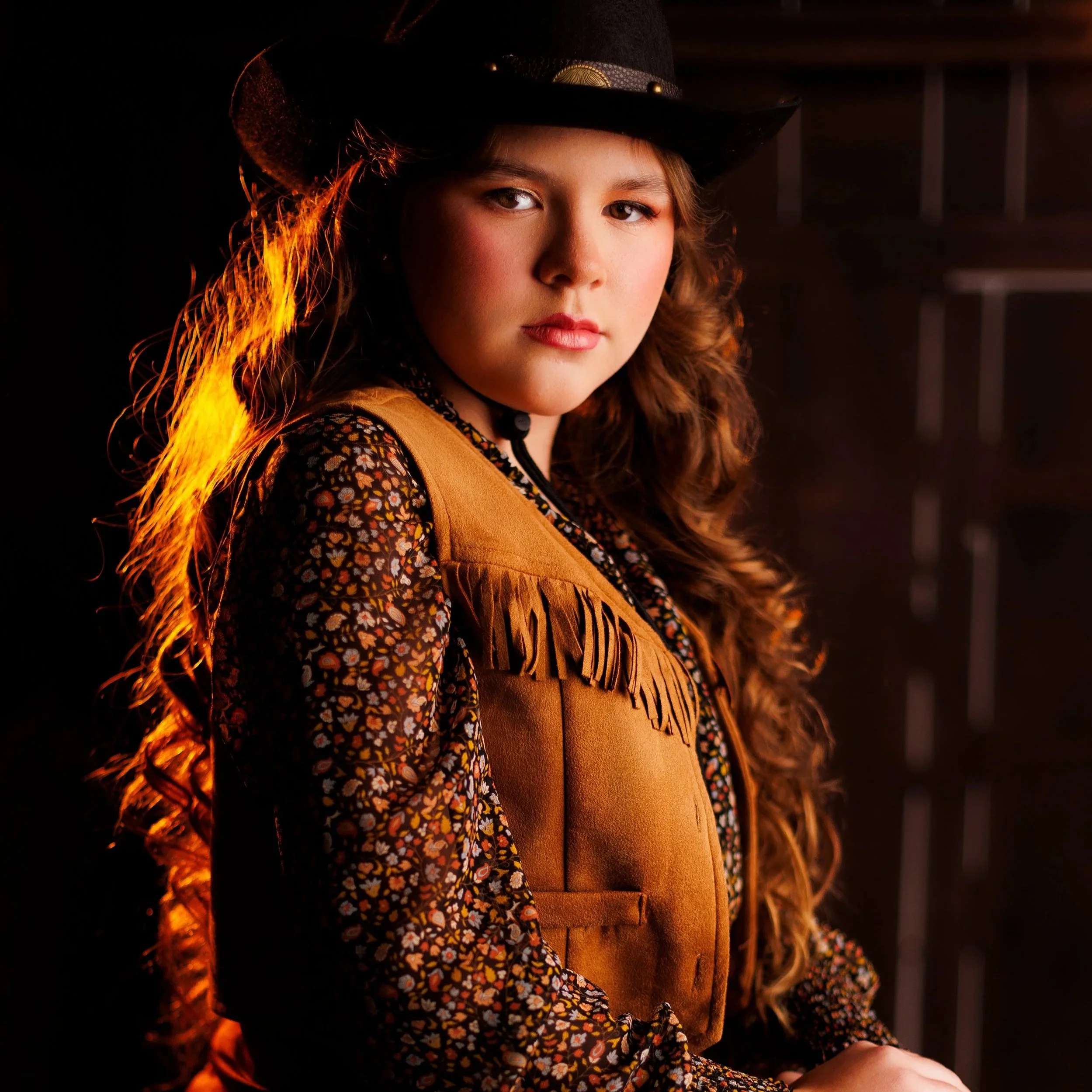A young woman dressed as a cowboy in a black hat and brown fringed vest with long curly hair, posing confidently indoors with a dark background.