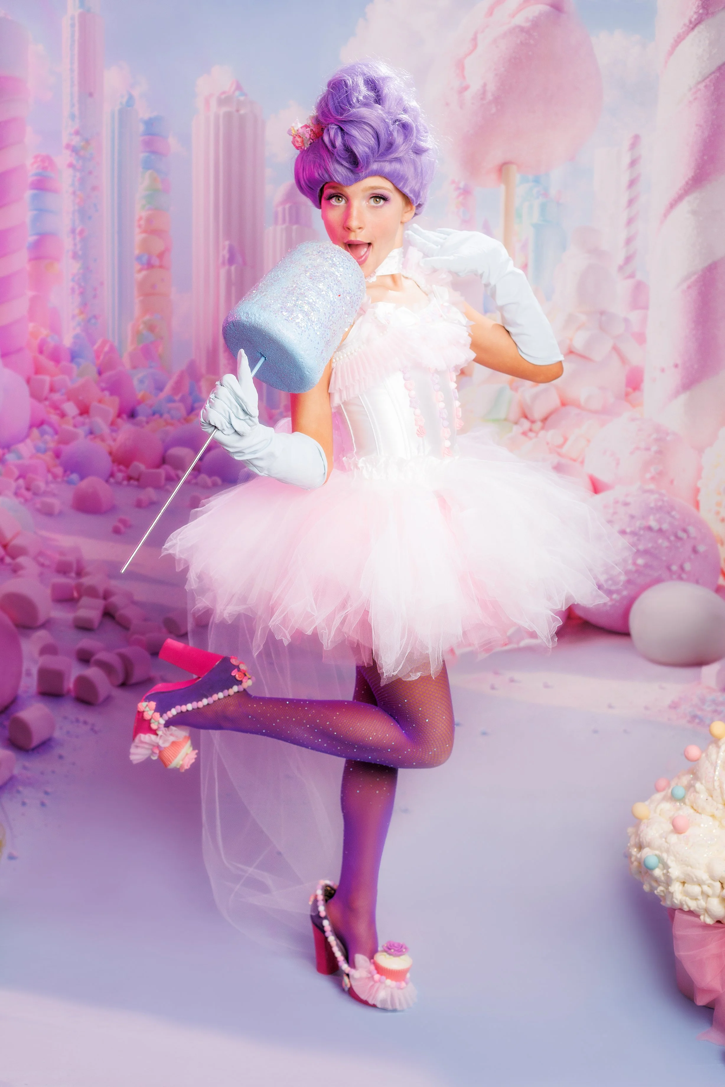 A woman dressed as a whimsical candy-themed character in a pastel pink and purple tutu, purple wig, and candy-themed accessories, holding a large glittery cotton candy stick in a fantasy-inspired candy land setting with pastel-colored decorations and