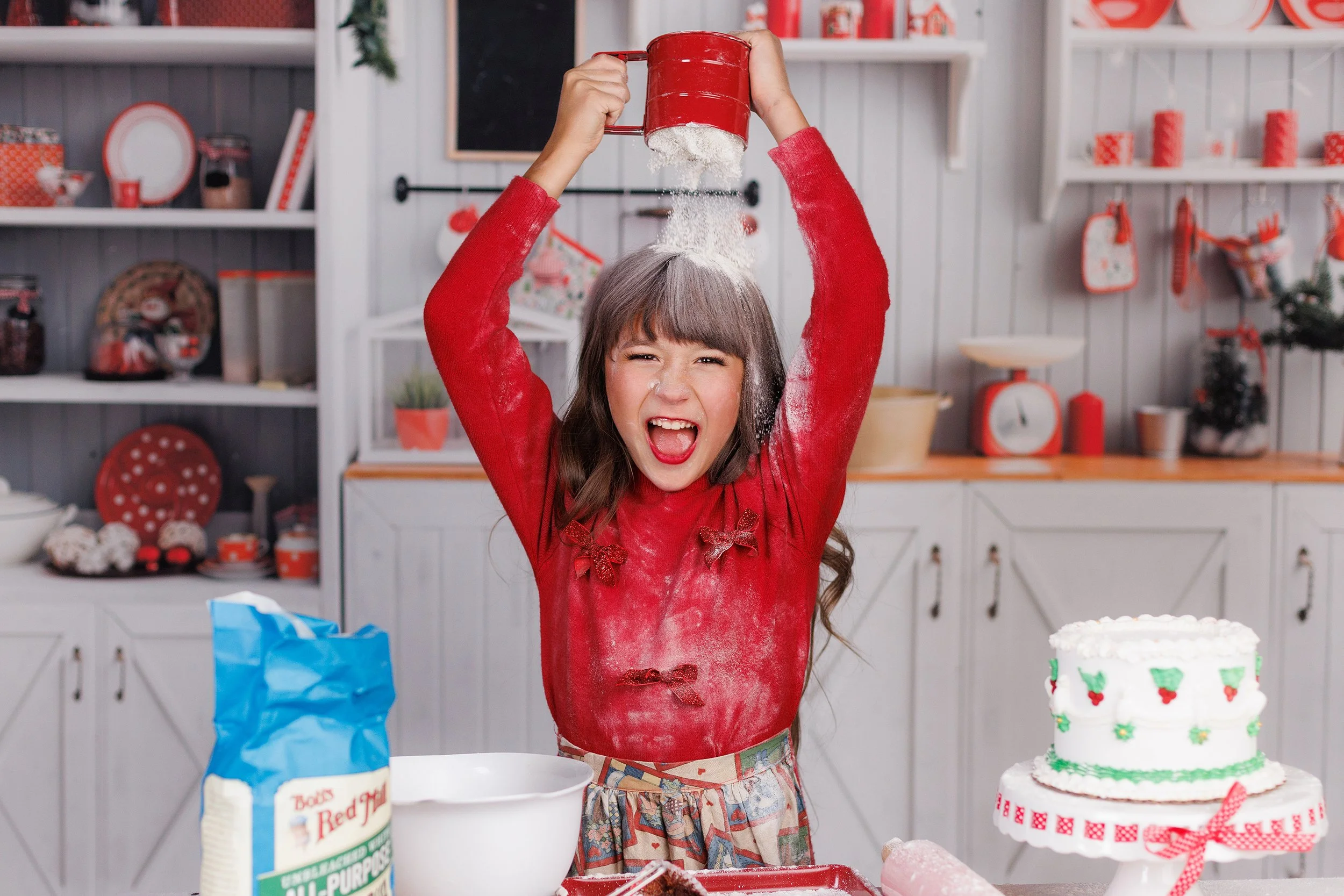 A girl in a red sweater is baking in a decorated kitchen. She is pouring flour over her head, with flour in the air. There is a cake decorated with holiday-themed decorations on the table.