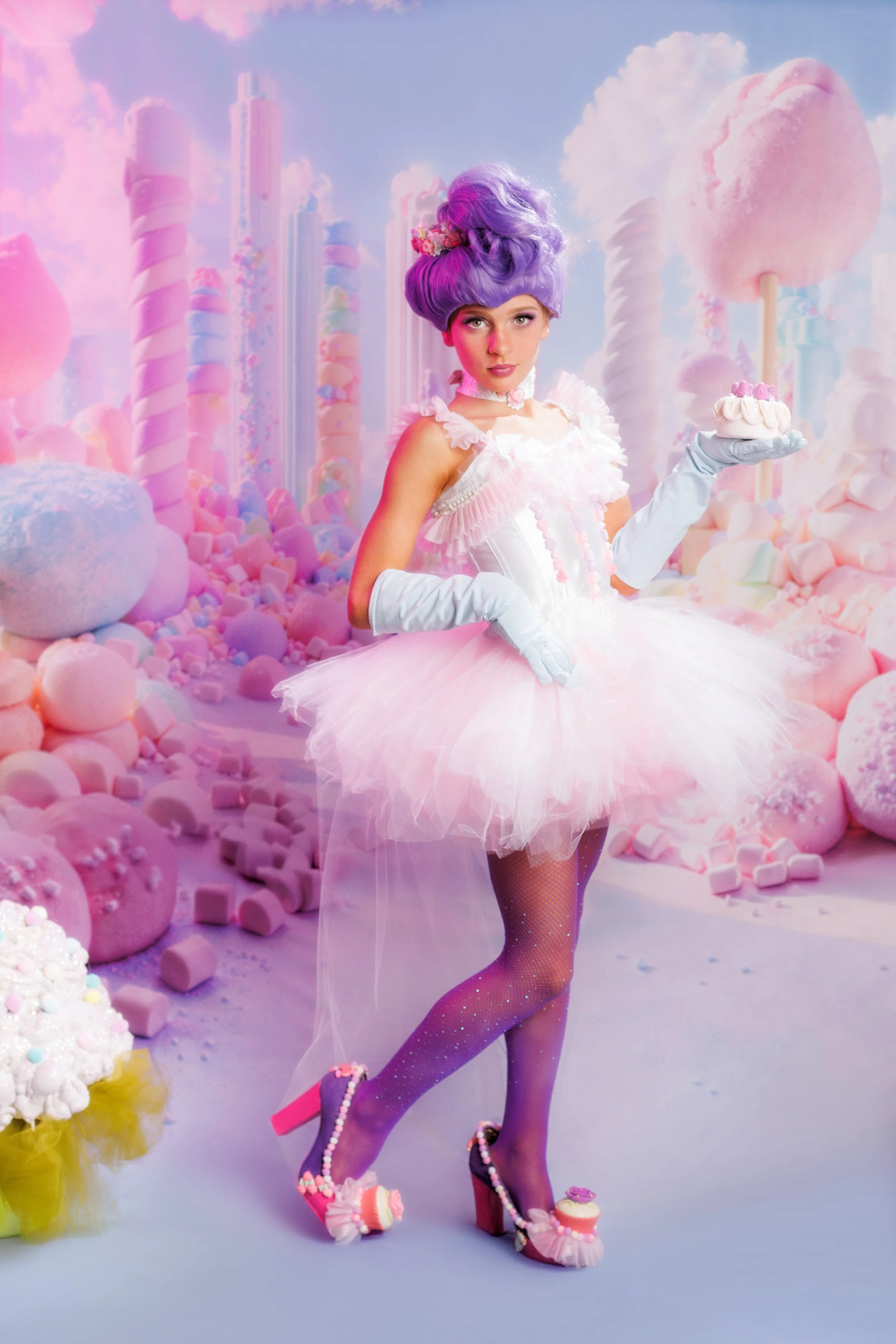 A young woman dressed in a pastel pink ballerina costume with a fluffy tutu, holding a small cake, standing in an environment filled with pink and white candy decorations and giant marshmallows.