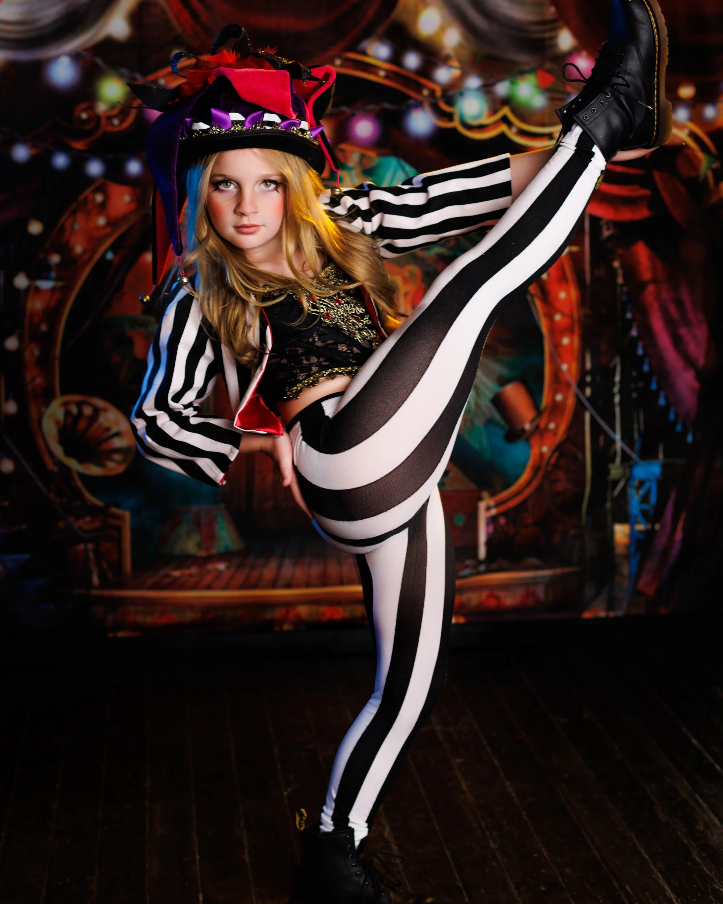 A young girl dressed as a circus performer or acrobat, wearing a colorful hat and striped black and white pants, poses with one leg raised high in front of a carnival or circus backdrop.