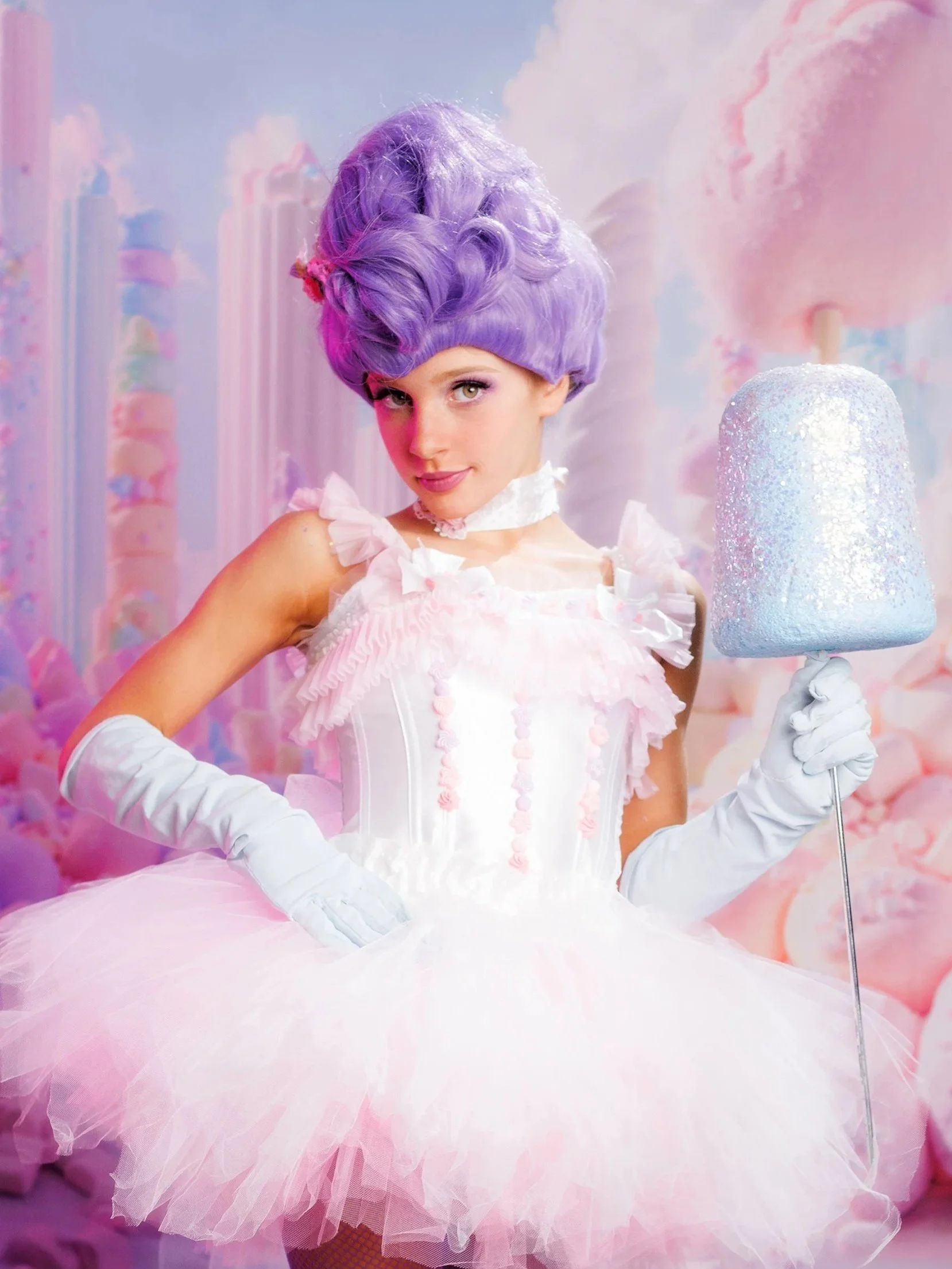 A woman dressed in a pink, princess-style tutu dress with ruffles, lace, and pink pom-poms, wearing long white gloves, holding a sparkly marshmallow on a stick, with a pastel-colored, candy-themed background.