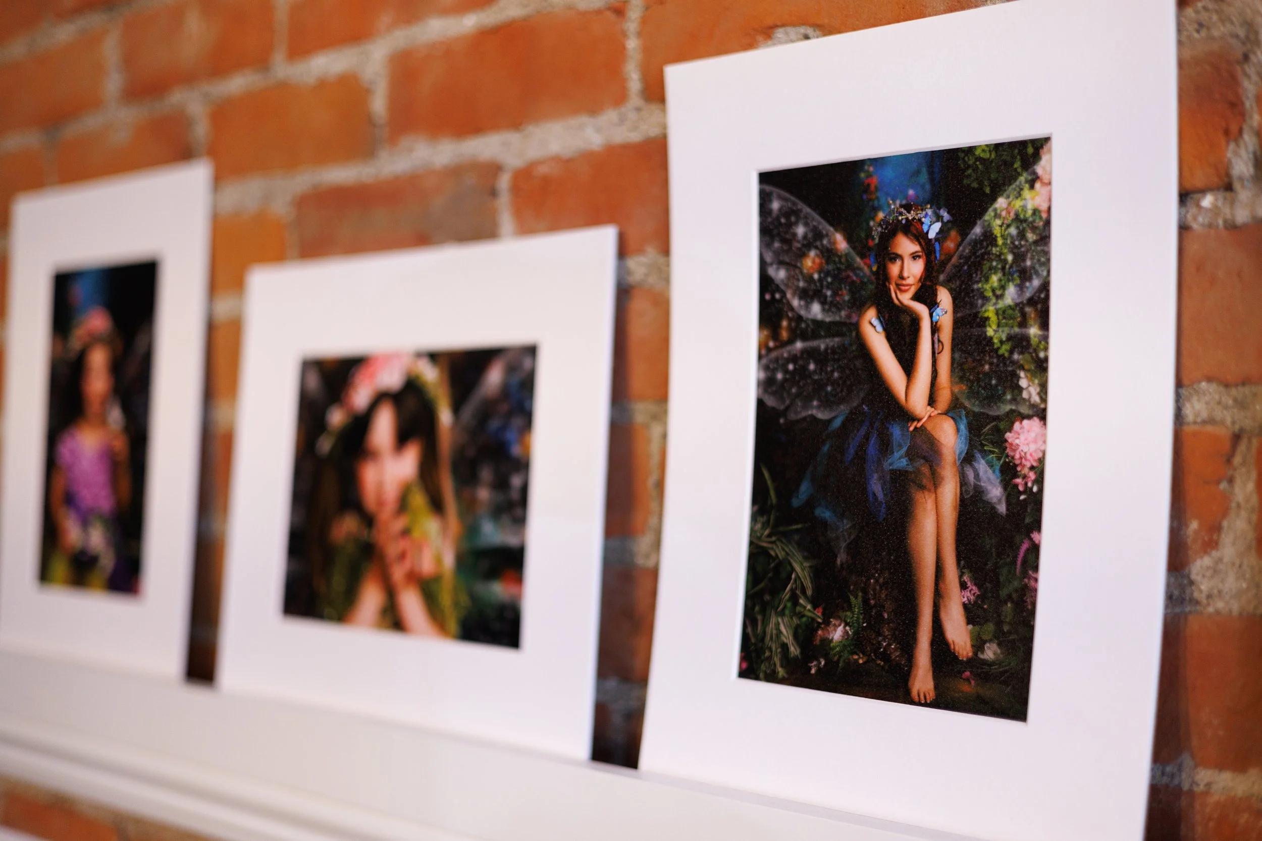 Fine art matted portrait prints displayed on a shelf against a brick wall, featuring whimsical fantasy-themed children’s portrait photography.