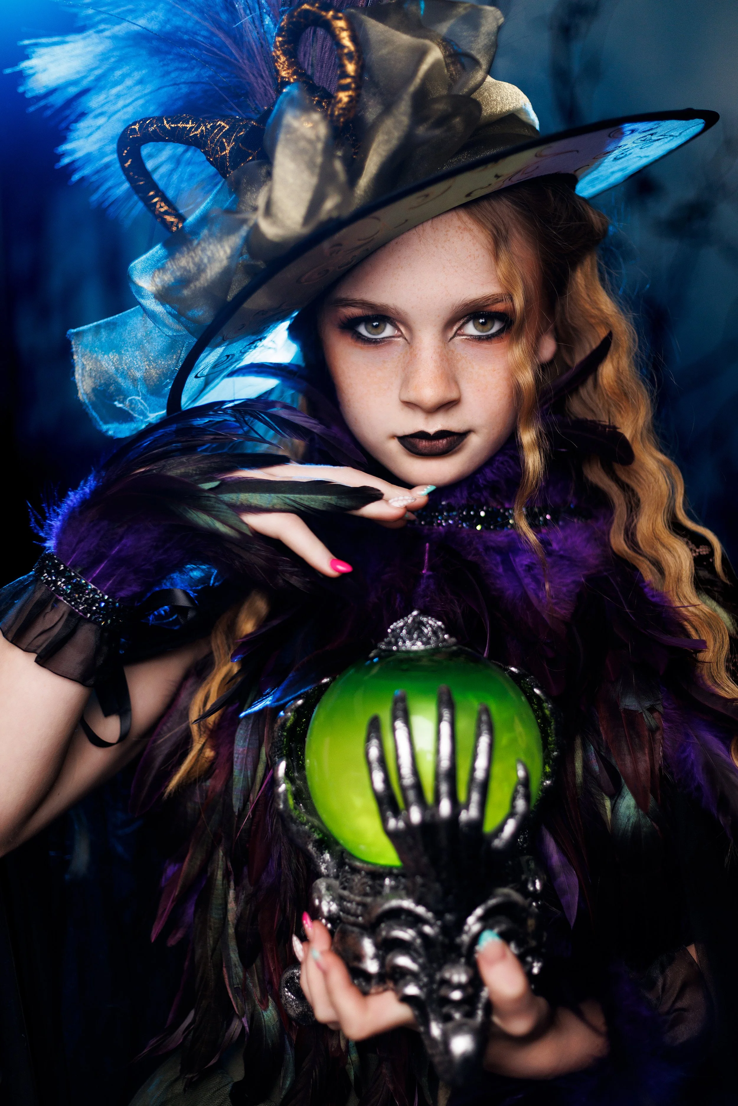 A person dressed as a witch with a large, elaborate hat adorned with feathers and ribbons, holding a green crystal ball with a decorative metallic skull base, dressed in dark, feathered clothing with bold makeup, dark lipstick, and long, curly hair.