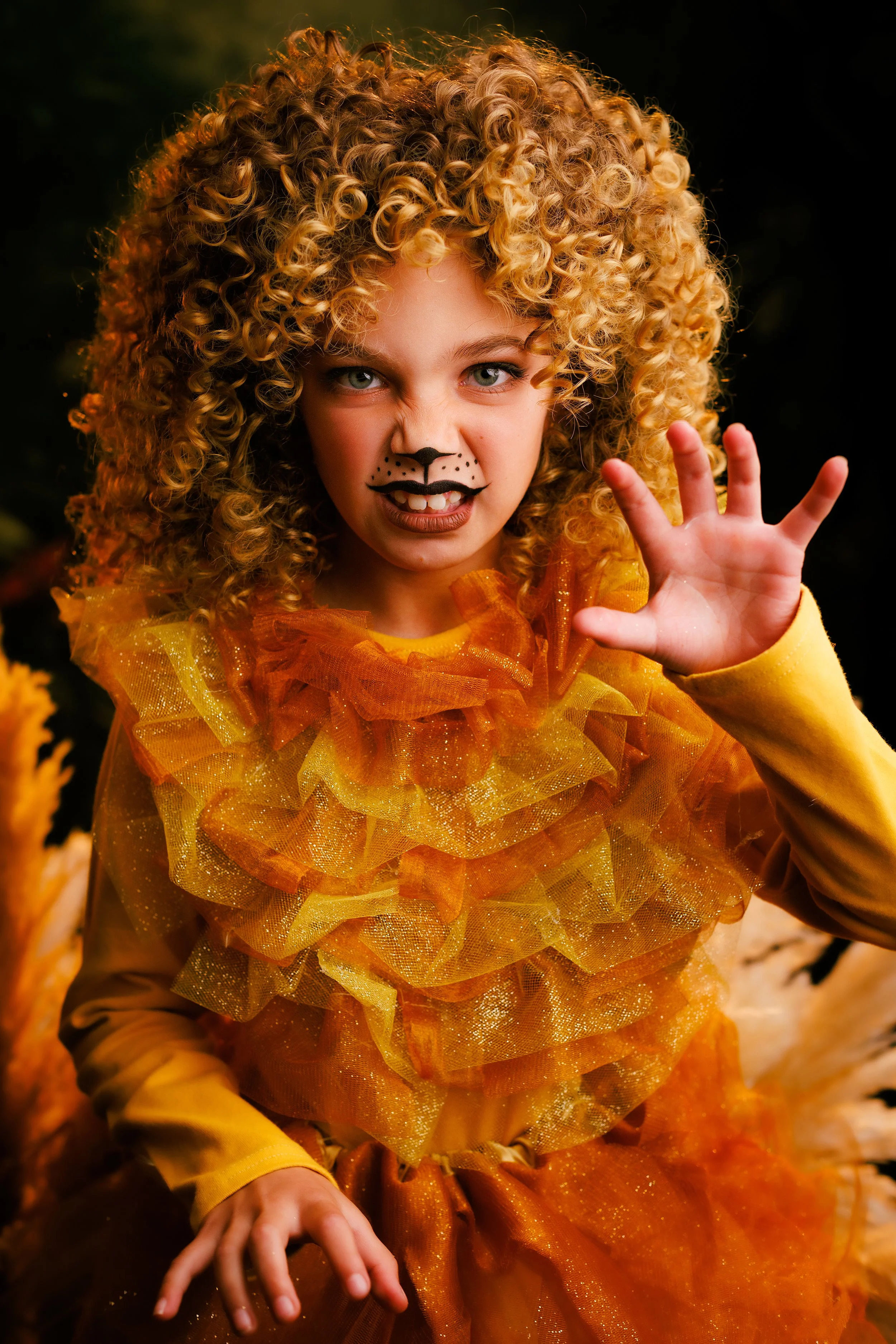 Young girl dressed in a lion costume with orange and yellow ruffled fabric, face painted with a lion nose and whiskers, making a fierce expression and reaching out with her hand.