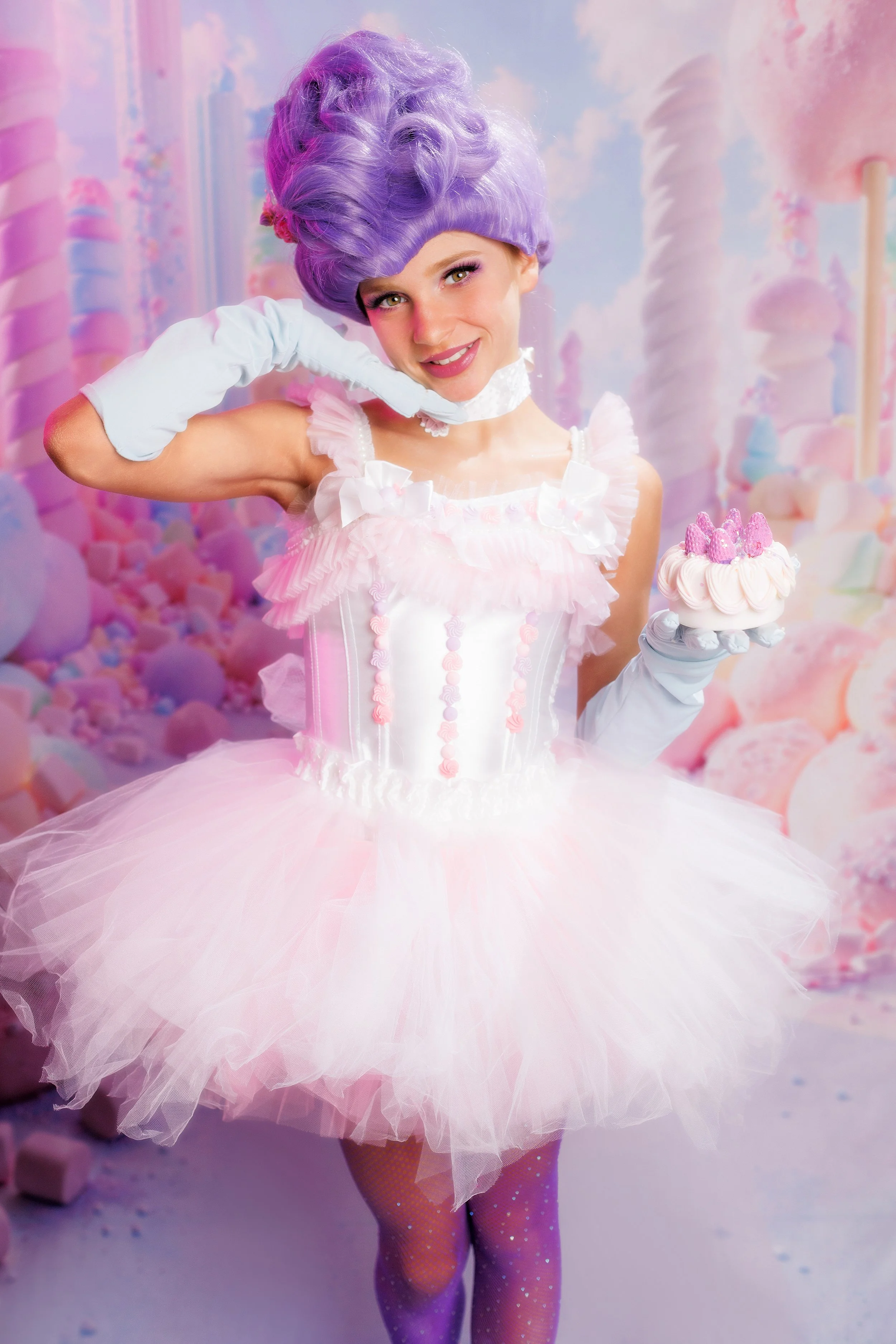 A woman dressed as a candy princess, with lavender hair styled in an elaborate updo, wearing a pink tutu and a white corset, holding a cupcake topped with pink berries, in a pastel candy-themed backdrop.
