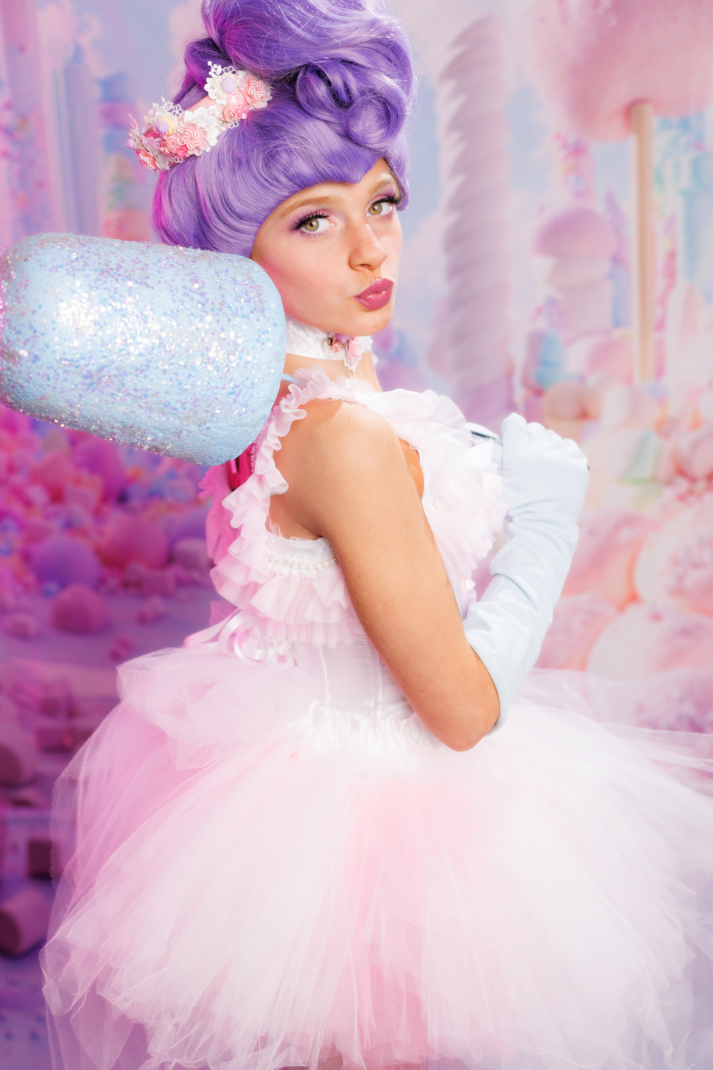 A woman with purple hair styled in an elaborate updo, wearing a pink tutu dress with white gloves and a crown, standing in a pastel-colored, candy-themed room.