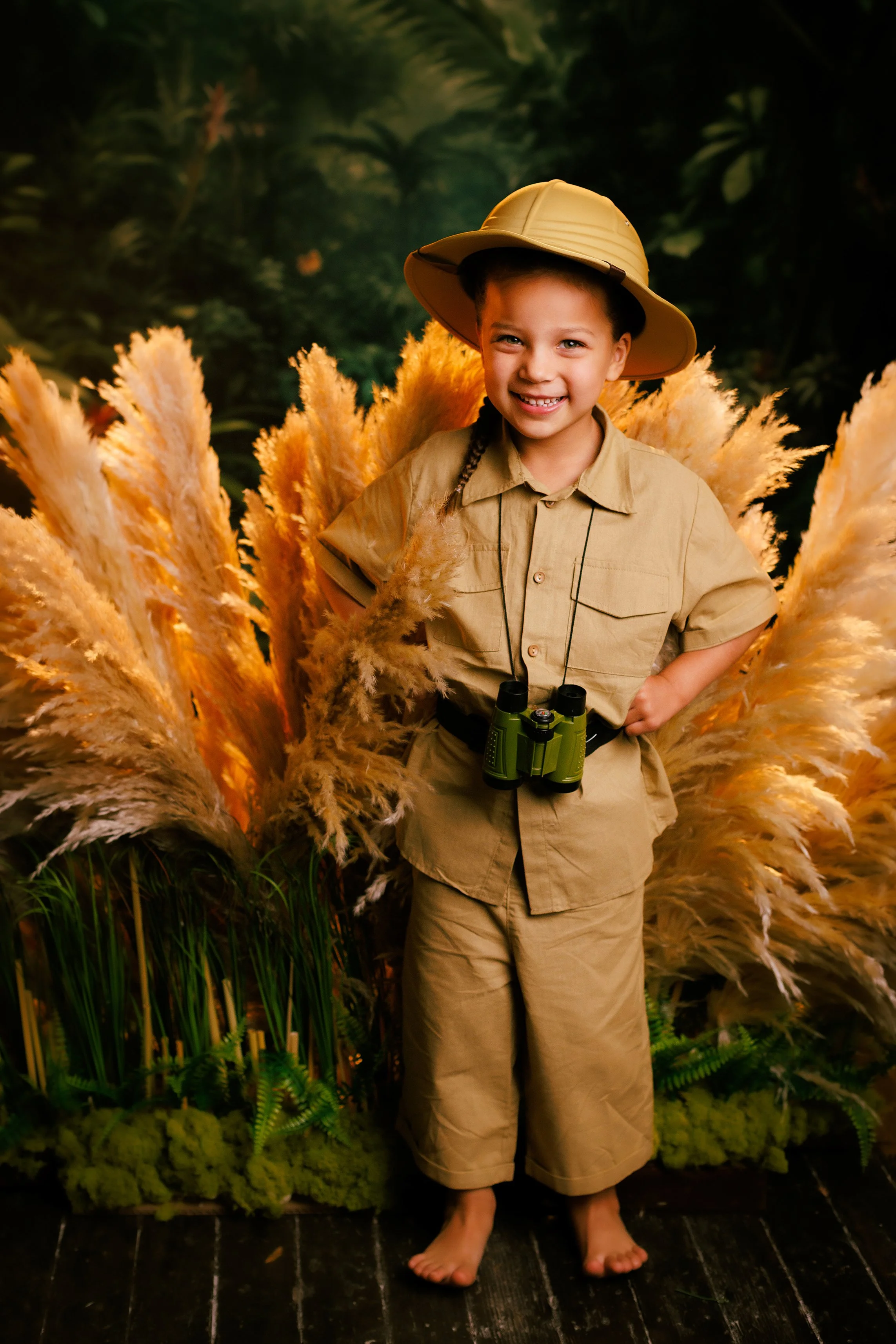 A young girl dressed in explorer safari attire, including a hat and khaki outfit, stands in front of a jungle backdrop with tall pampas grass and greenery, smiling at the camera with binoculars hanging around her neck.