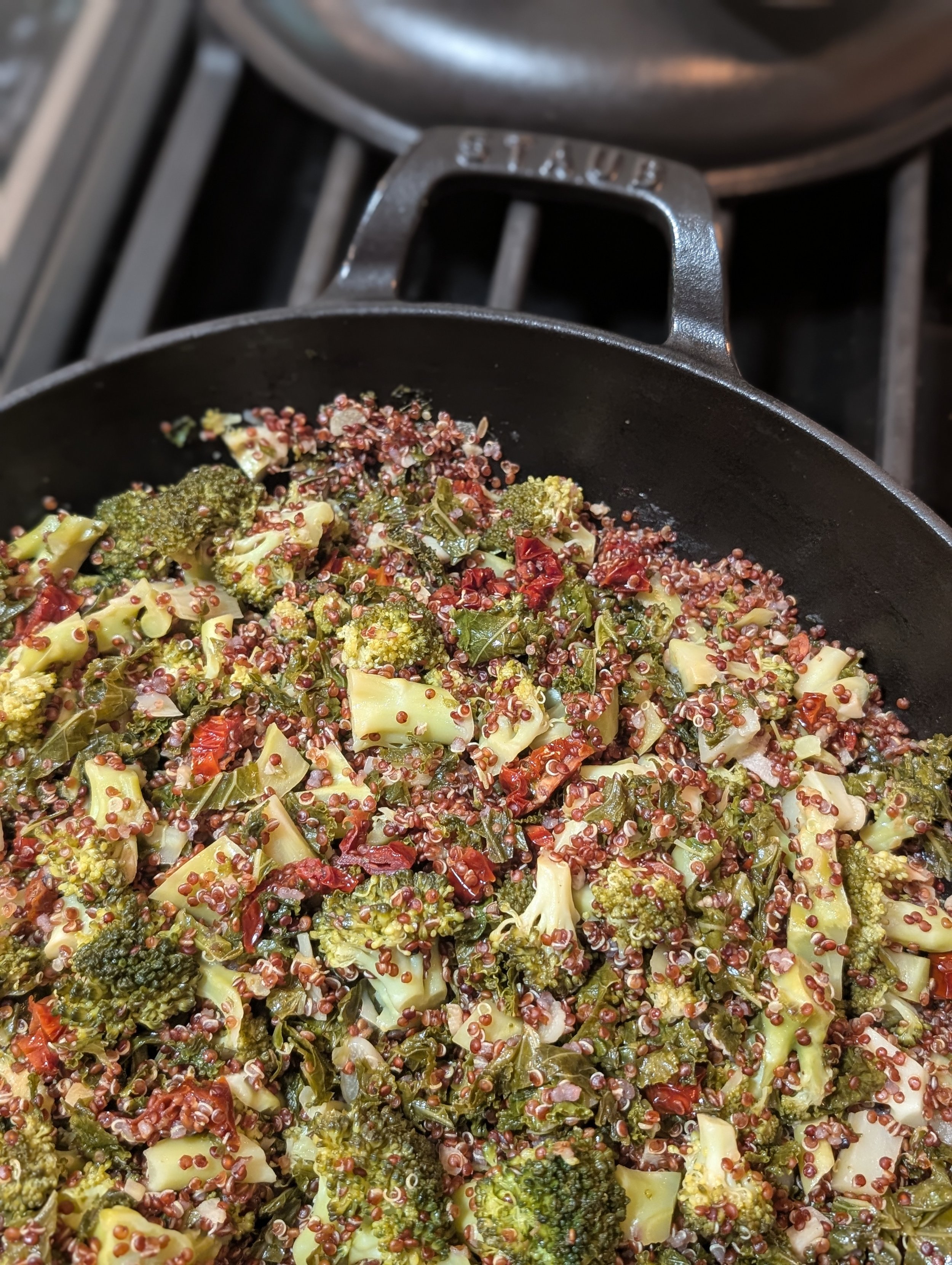 A cast iron Staub braiser holds braised broccoli and kale with red quinoa, sundried tomatoes and garlic.