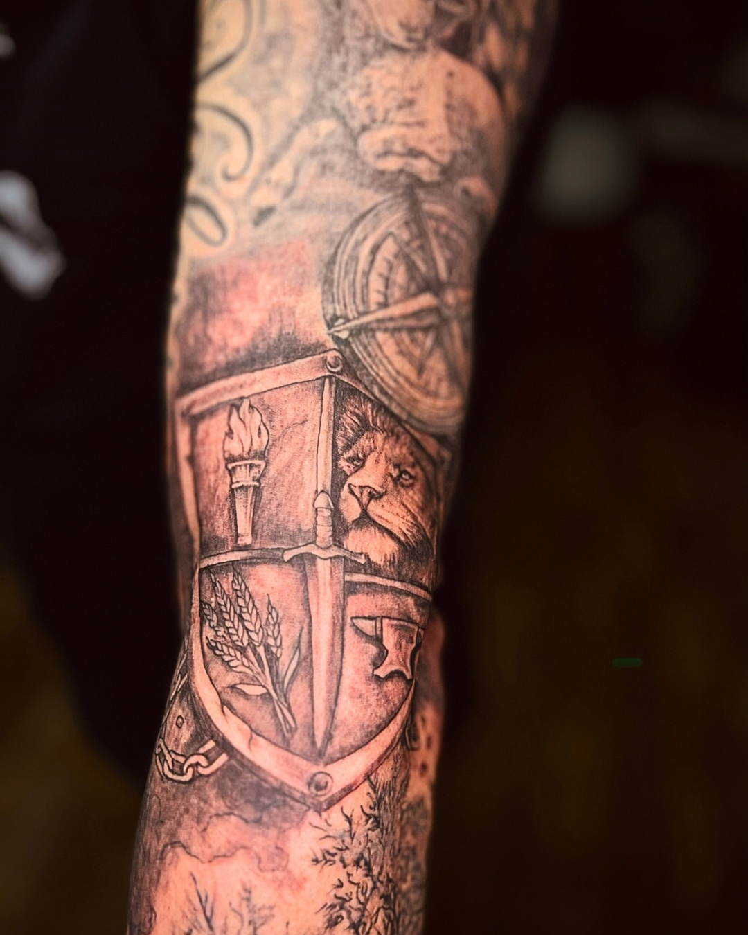 Close-up of a custom black and grey family crest tattoo featuring a lion, sword, torch, wheat, and heraldic shield symbolizing courage, honor, wisdom, and legacy, professionally tattooed at Brass Knuckles Ink in the Bronx.