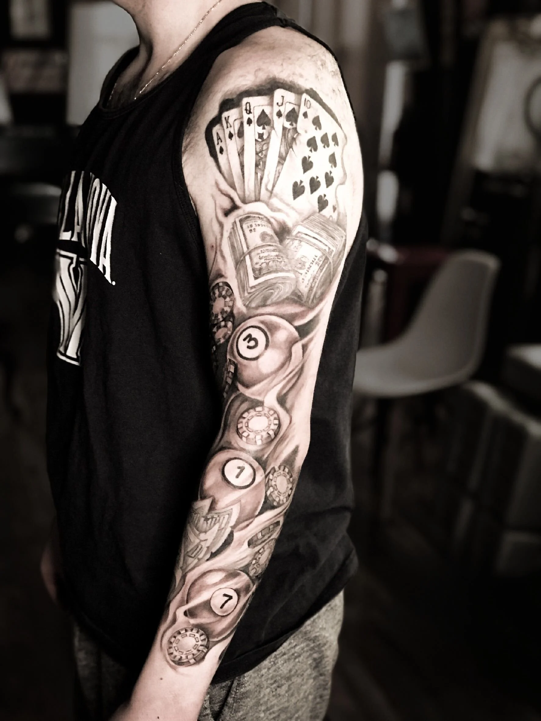 Black and grey gambling themed sleeve tattoo featuring playing cards, billiard balls, and casino elements on upper arm in Bronx NYC.