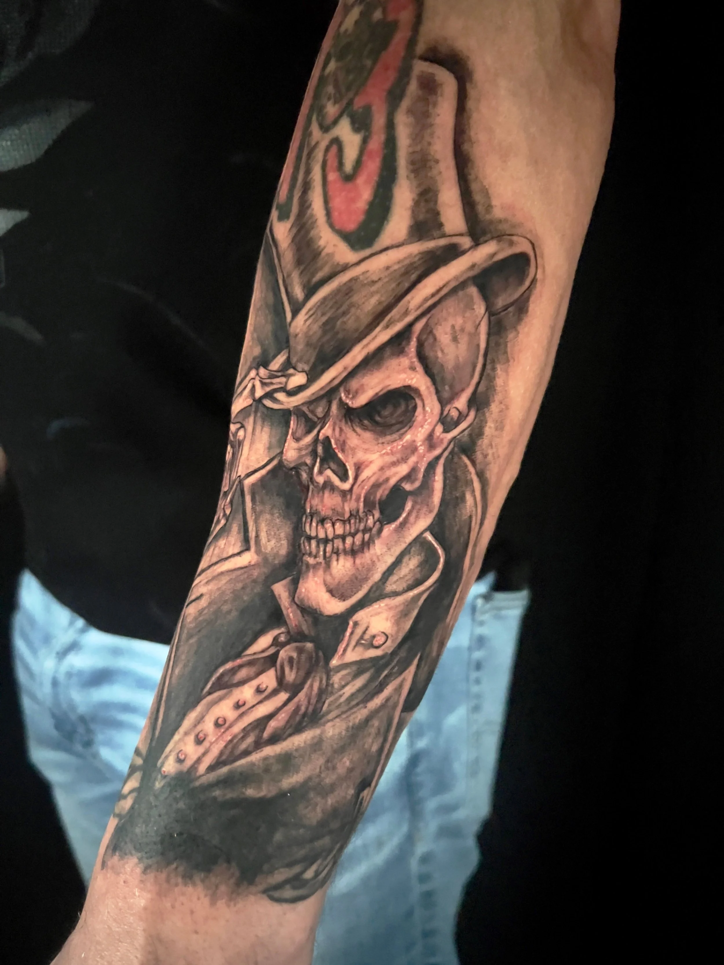 This black and grey gangster skull forearm tattoo features a sharply detailed skull wearing a fedora and suit, inspired by classic Chicano realism. Smooth gradient shading and bold contrast create depth and dimension throughout the piece
