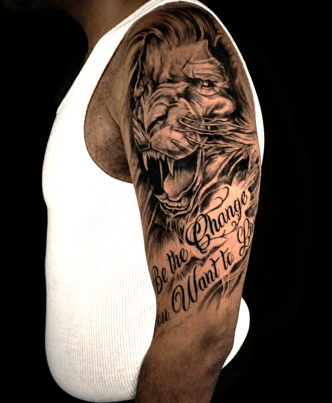 Custom black and grey lion portrait realism tattoo on the upper arm and shoulder created at Brass Knuckles Ink Tattoo Studio in the Bronx, NYC, featuring detailed realism, flowing script lettering, and custom sleeve composition.