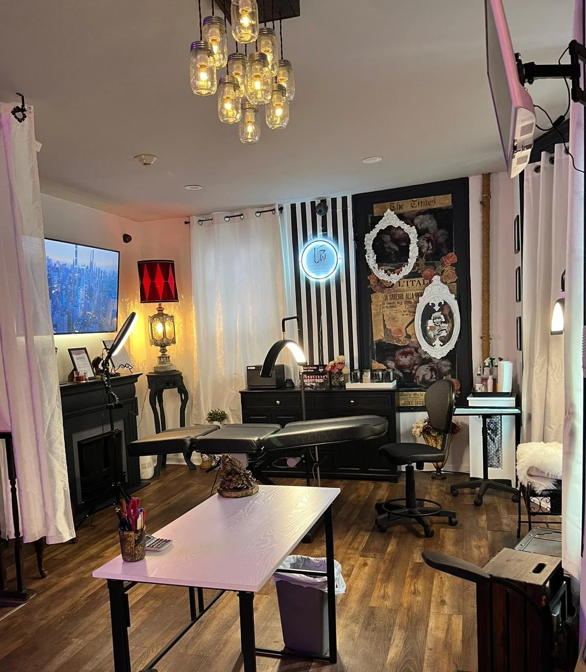 A well-decorated massage therapy room with a massage table, black furniture, and various decorative items. The room has wooden floors, white curtains, and soft lighting. Luxury Permanent Makeup studio in NYC metro area
