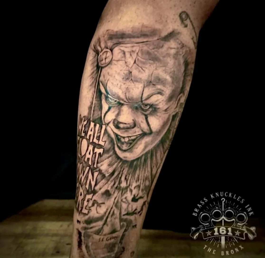 This black and grey realism tattoo features Pennywise the clown from the movie IT, captured with a dark, sinister expression and fine realistic detail. The design emphasizes textured shading, dramatic contrast, and lifelike facial features to create 