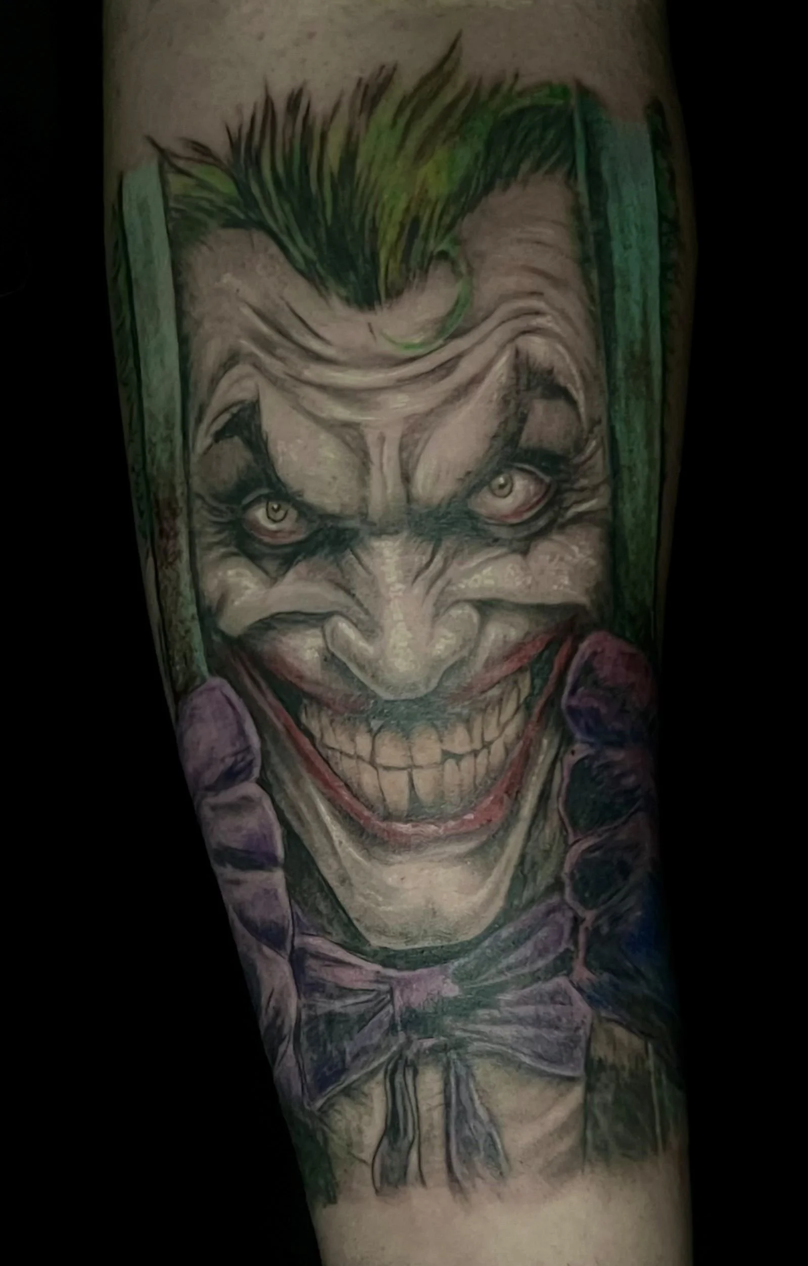 A tattoo of the Joker clown character with green hair, pale face, dark eye makeup, and a wide, toothy grin, wearing a purple suit with a bow tie.