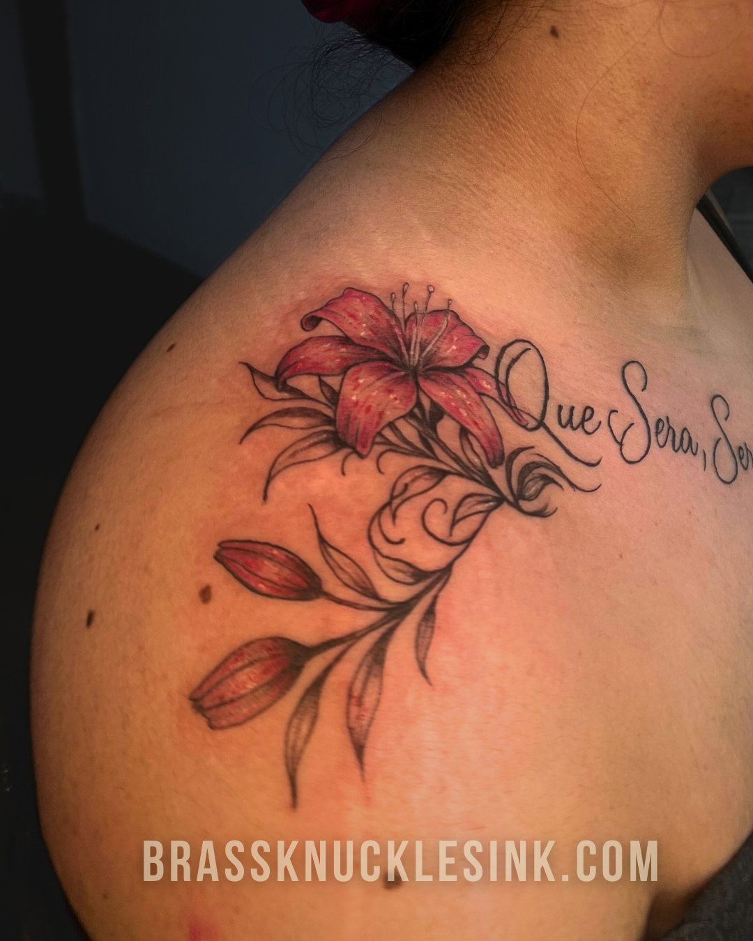 Custom shoulder tattoo of red lily and 'Que Sera, Sera' script from Brass Knuckles Ink – expert tattoos in a clean, comfortable studio.