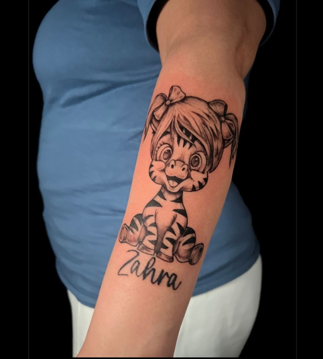 Black and grey anime-inspired zebra girl tattoo with bows and name lettering on the forearm by a Bronx tattoo artist. Focus keywords: Black and grey tattoo, anime-inspired tattoo, tiger girl, Bronx tattoo artist.