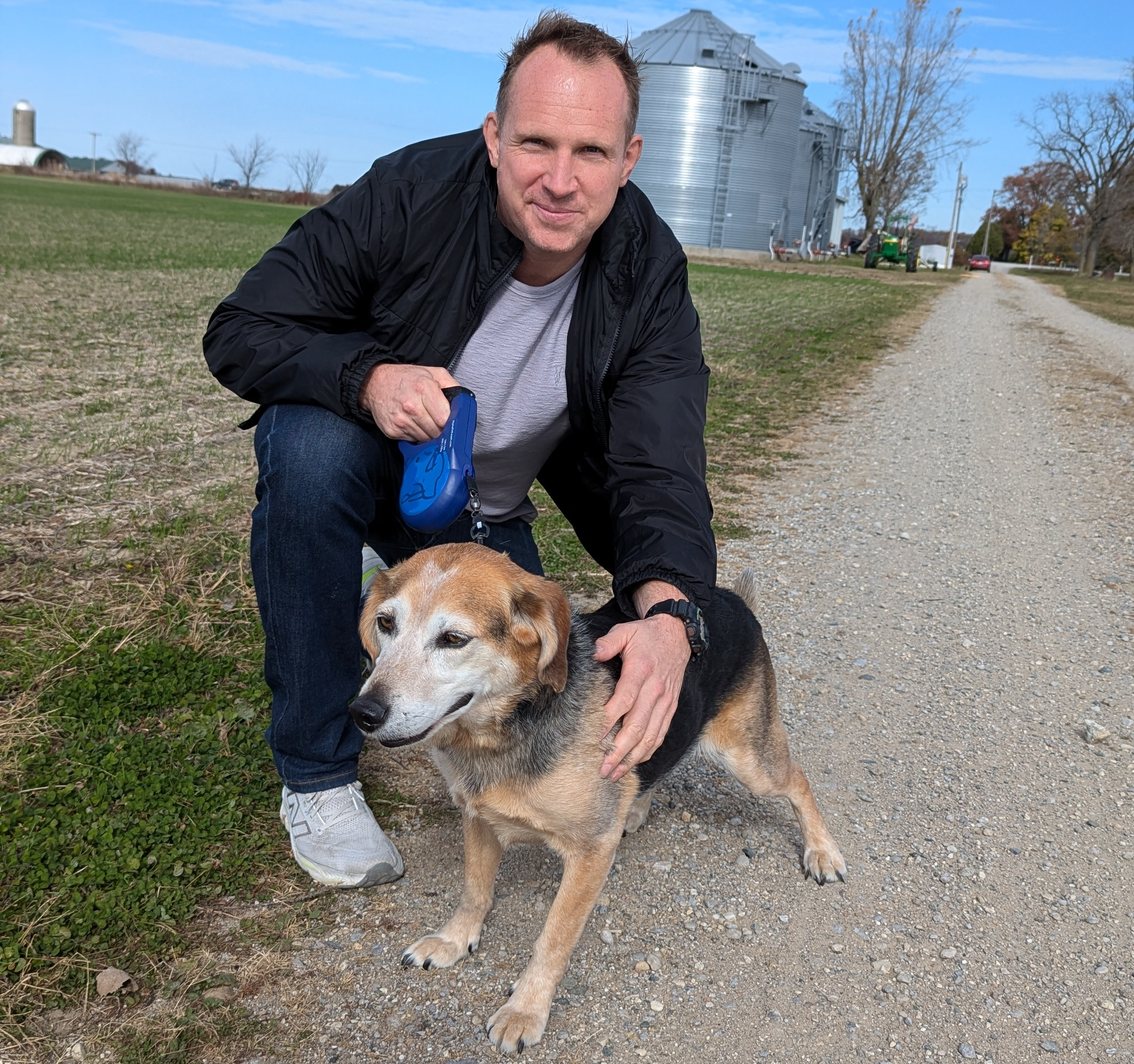 The inventor of the Alpha Leash crouching down outdoors on a dirt and grass path with his hand on a medium sized, tan and black dog and holding an Alpha Leash.