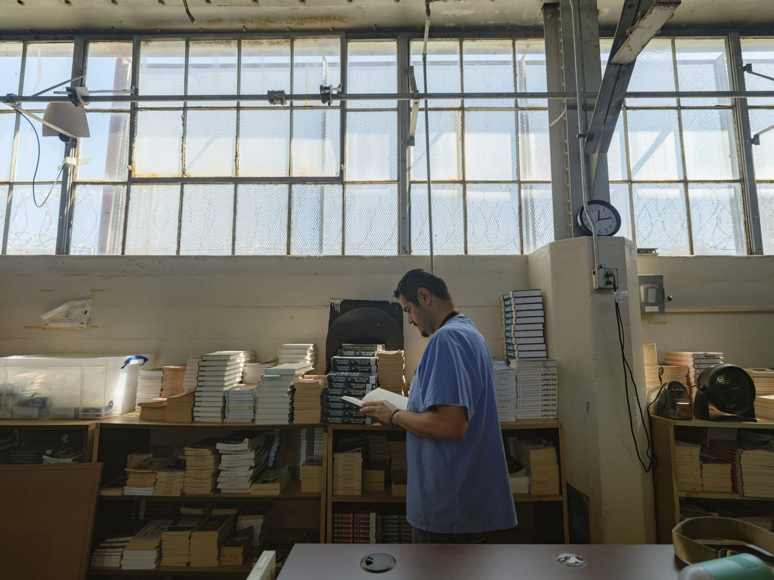 A man in a blue shirt stands in front of a table filled with stacked books inside a room with large windows and a clock on the wall.
