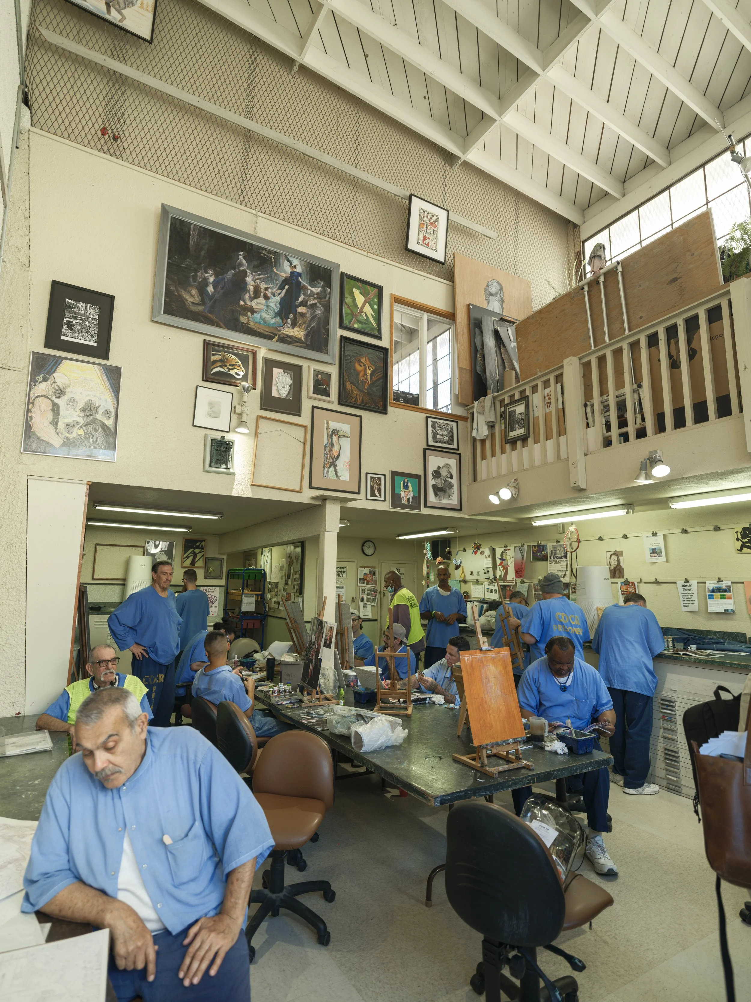 Group of people working on art projects and painting in an art studio with various artworks displayed on walls and a high ceiling.