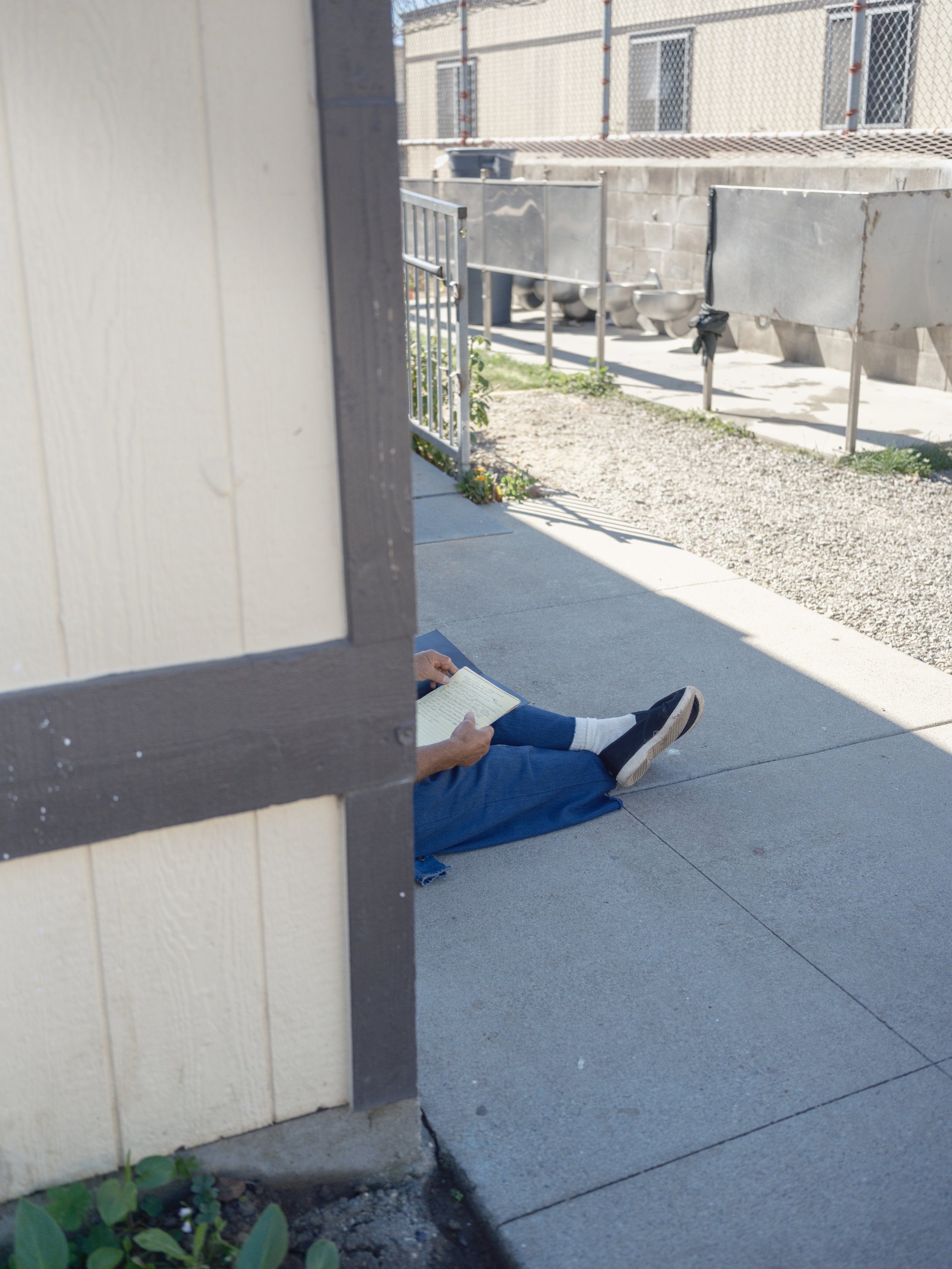 Person sitting on the sidewalk reading a book, seen partially from behind and from the side, with legs stretched out, wearing blue jeans and black shoes, in an outdoor urban setting with buildings, a chain-link fence, and some plants nearby.