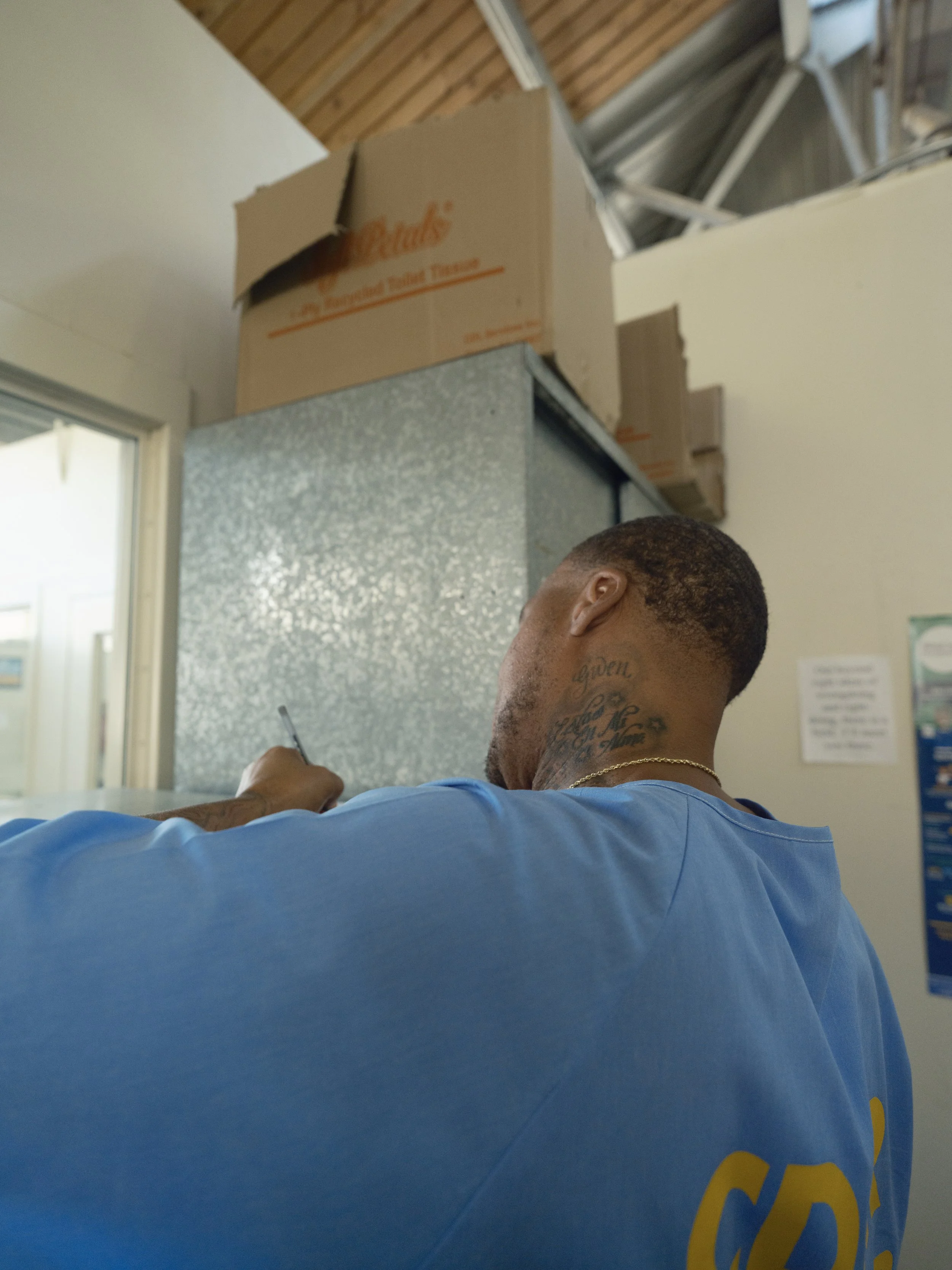 A man with tattoos on his neck wearing a light blue shirt, writing or drawing with a pen in a room with a wooden ceiling, partial window, and cardboard boxes on top of a metallic surface in the background.