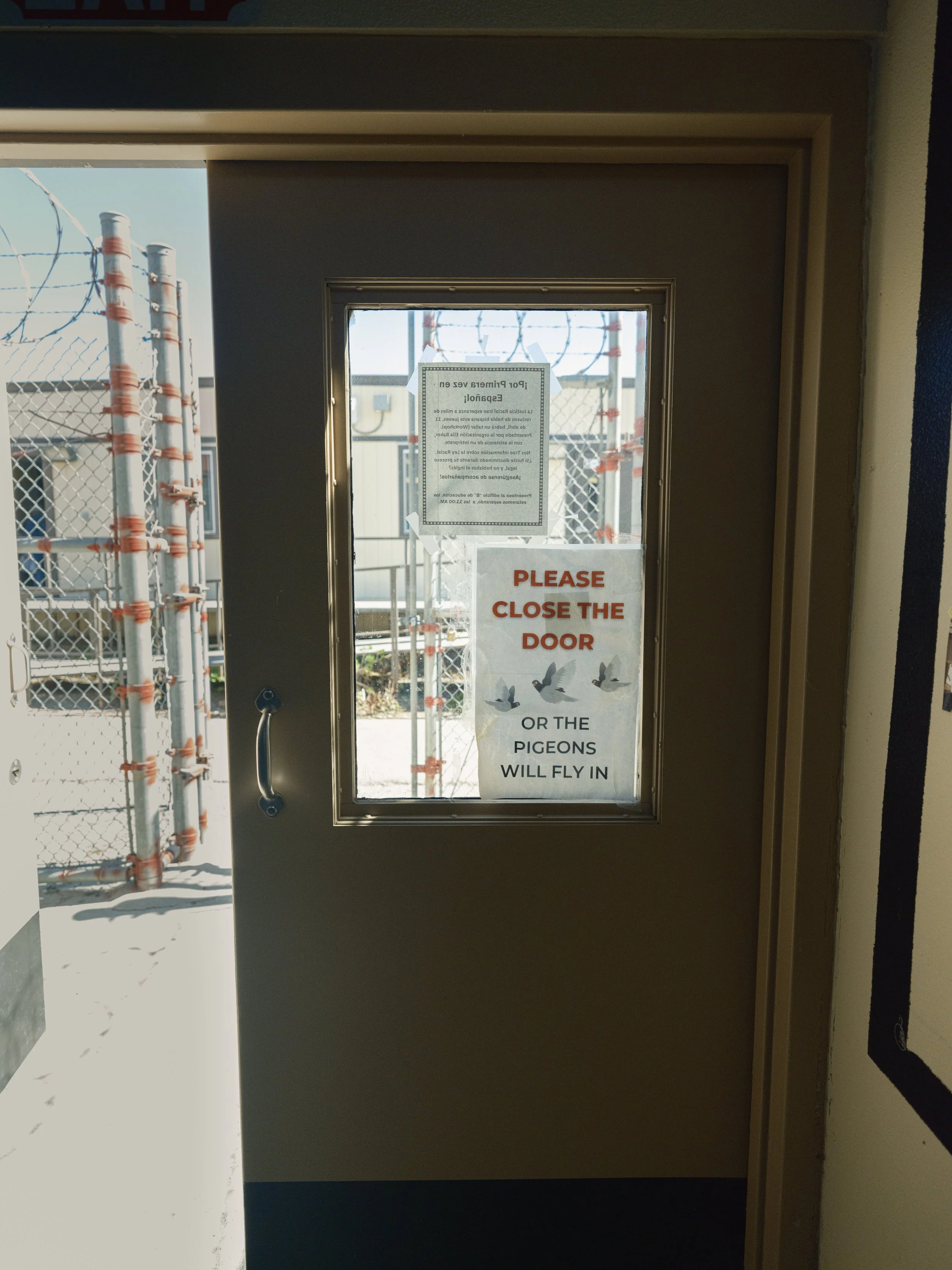 A door with a glass window featuring a sign that reads, 'Please close the door or the pigeons will fly in.' Outside the door, there is a chain-link fence with orange and white striped poles and barbed wire on top.