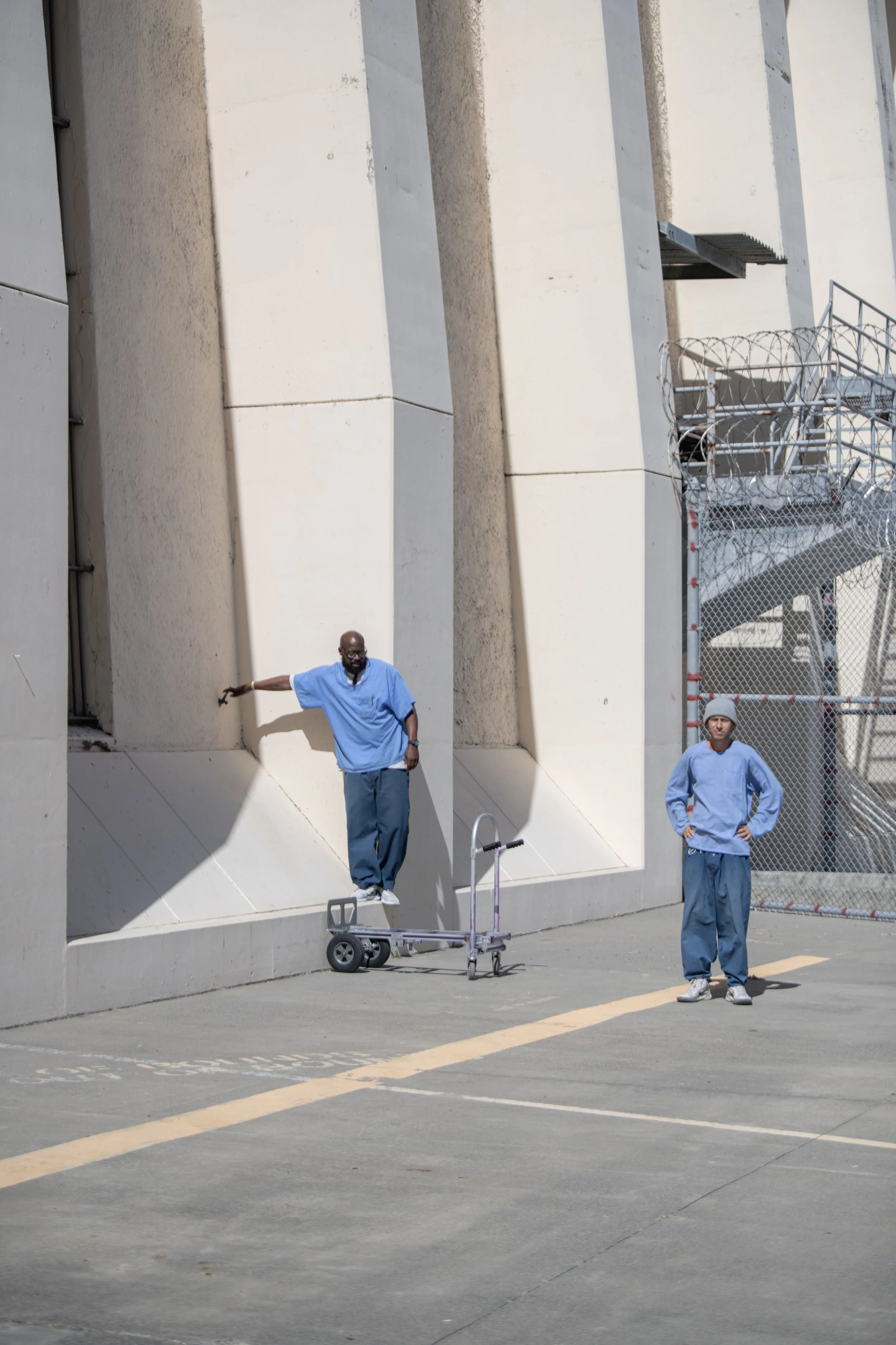 Two men in blue prison uniforms standing outside near a beige building. One man is raising his arm while the other stands with his hands in his pockets. There is a small cart with wheels in front of the man with his arm raised. A chain-link fence and