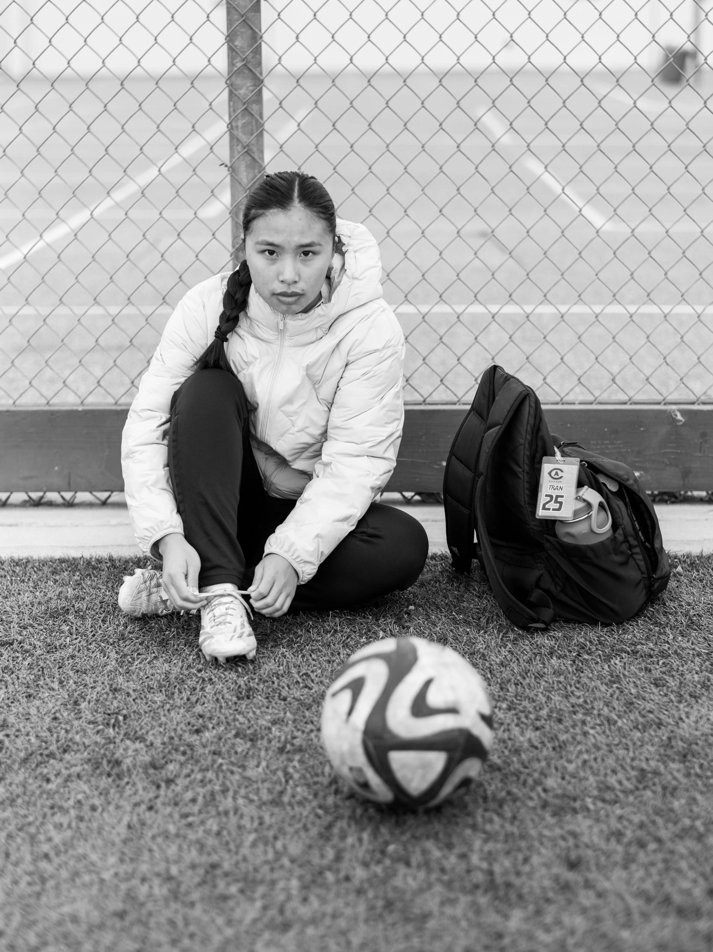 A young woman in athletic clothing and a braided hairstyle is sitting on the ground in front of a chain-link fence with a tennis court in the background. She is tying her shoelaces next to her backpack and a tennis ball.