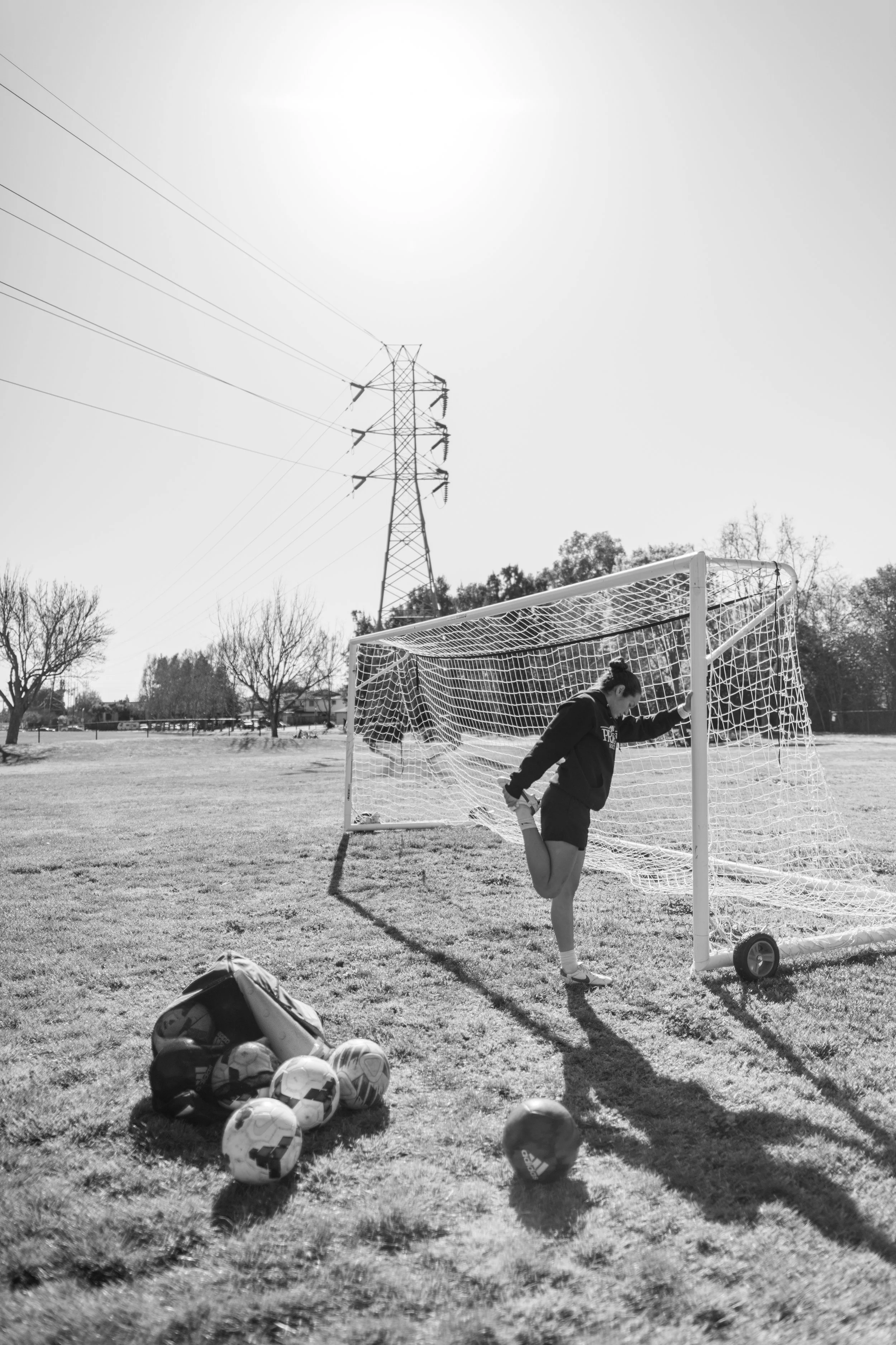 A girl in athletic clothing practicing soccer near a goalpost on a sunny day. Soccer balls are scattered on the grass, and there are leafless trees and the power lines in the background.