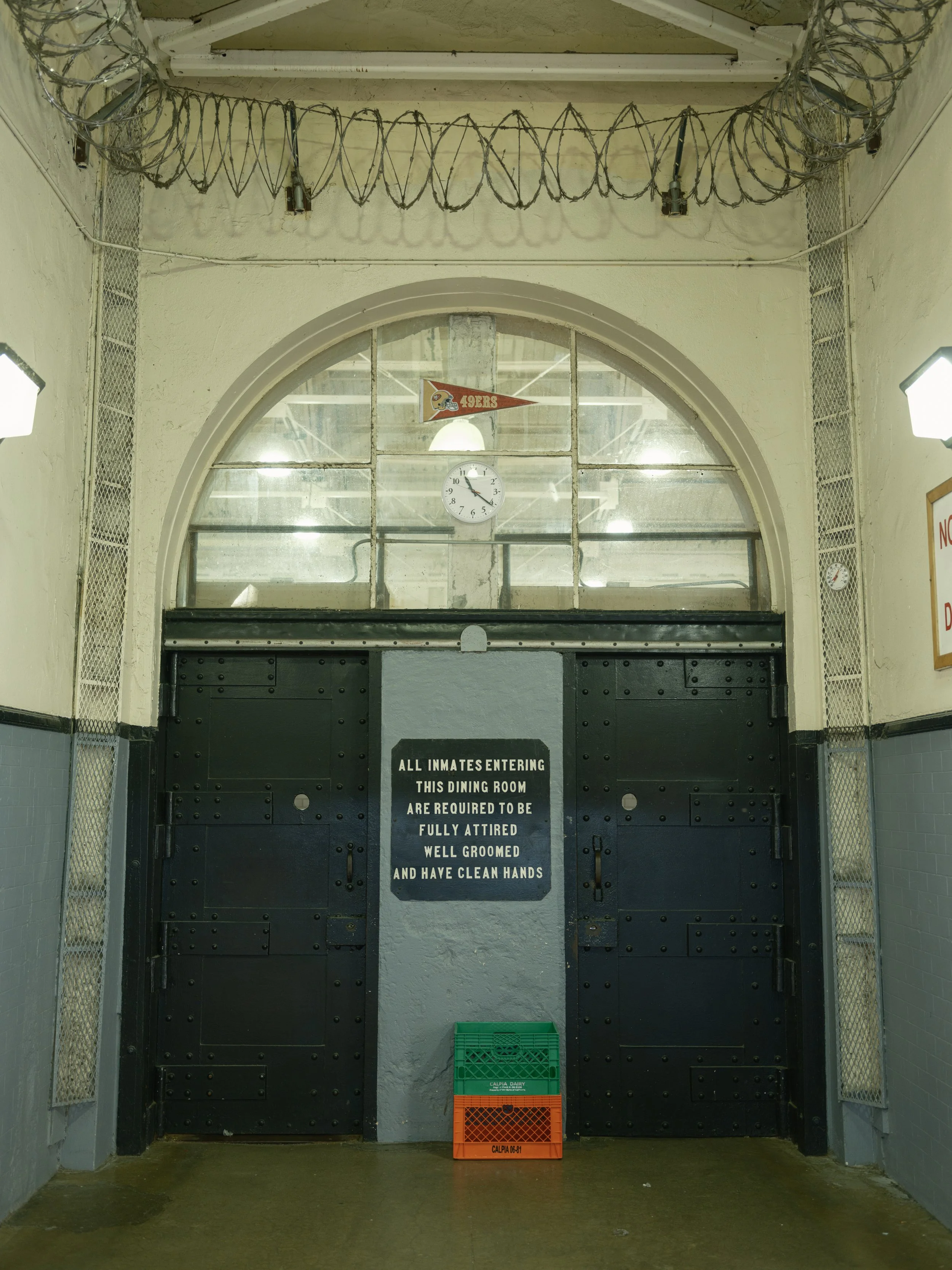 Black metal gate with a sign that reads: All inmates entering this dining room are required to be fully attired, well groomed, and have clean hands. Two plastic crates, one green and one orange, are placed in front of the gate. There is a large windo