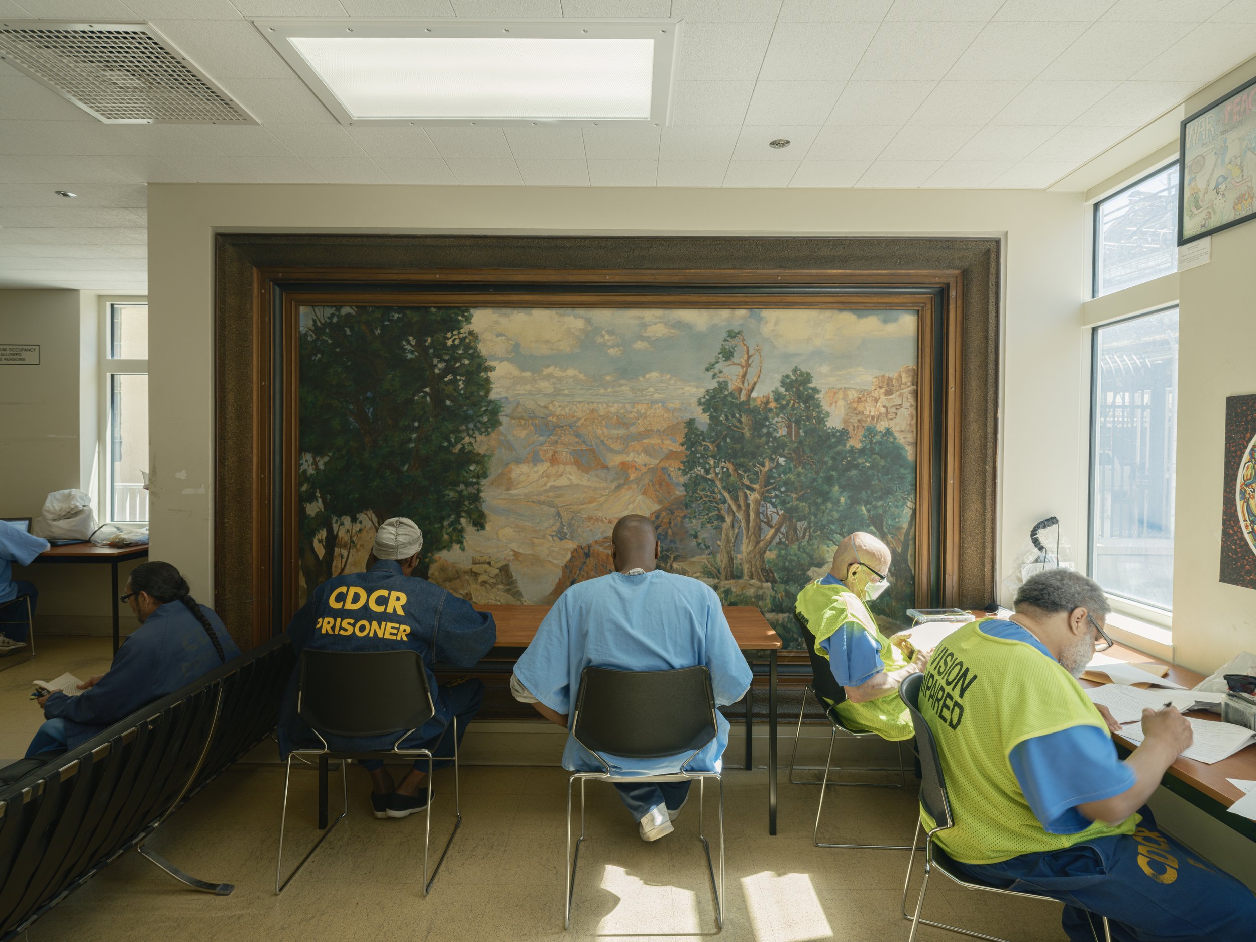 Group of incarcerated individuals sitting at tables, some wearing blue uniforms, in an indoor setting with a large landscape painting on the wall behind them and windows on the side. Some are engaged in paperwork, others are wearing masks.