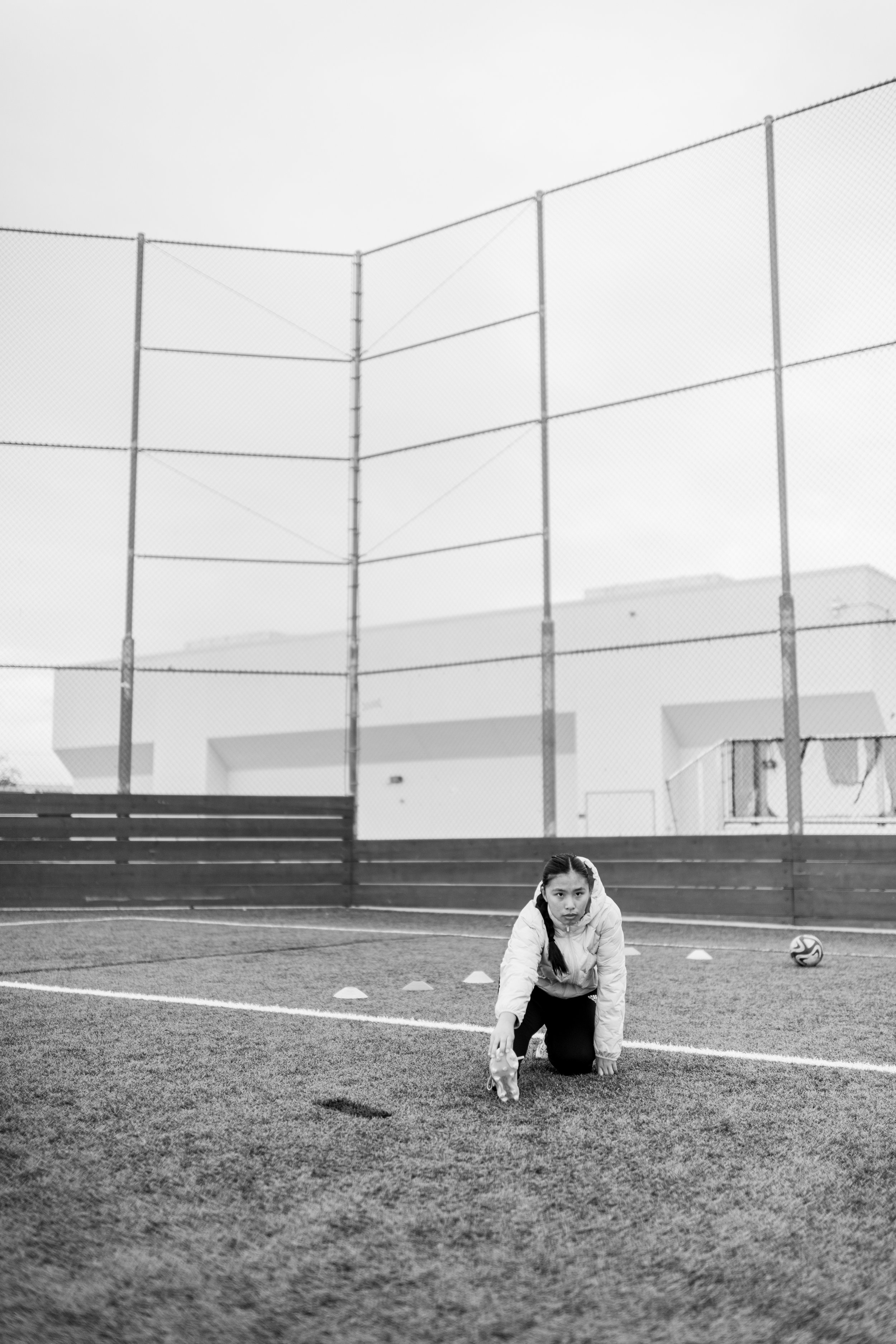 A young girl is in a sports field, kneeling on the ground, with one knee on the turf and one foot on the ground, stretching or warming up. She is wearing a light-colored jacket and has dark hair in braids. A soccer ball and small cones are on the fie