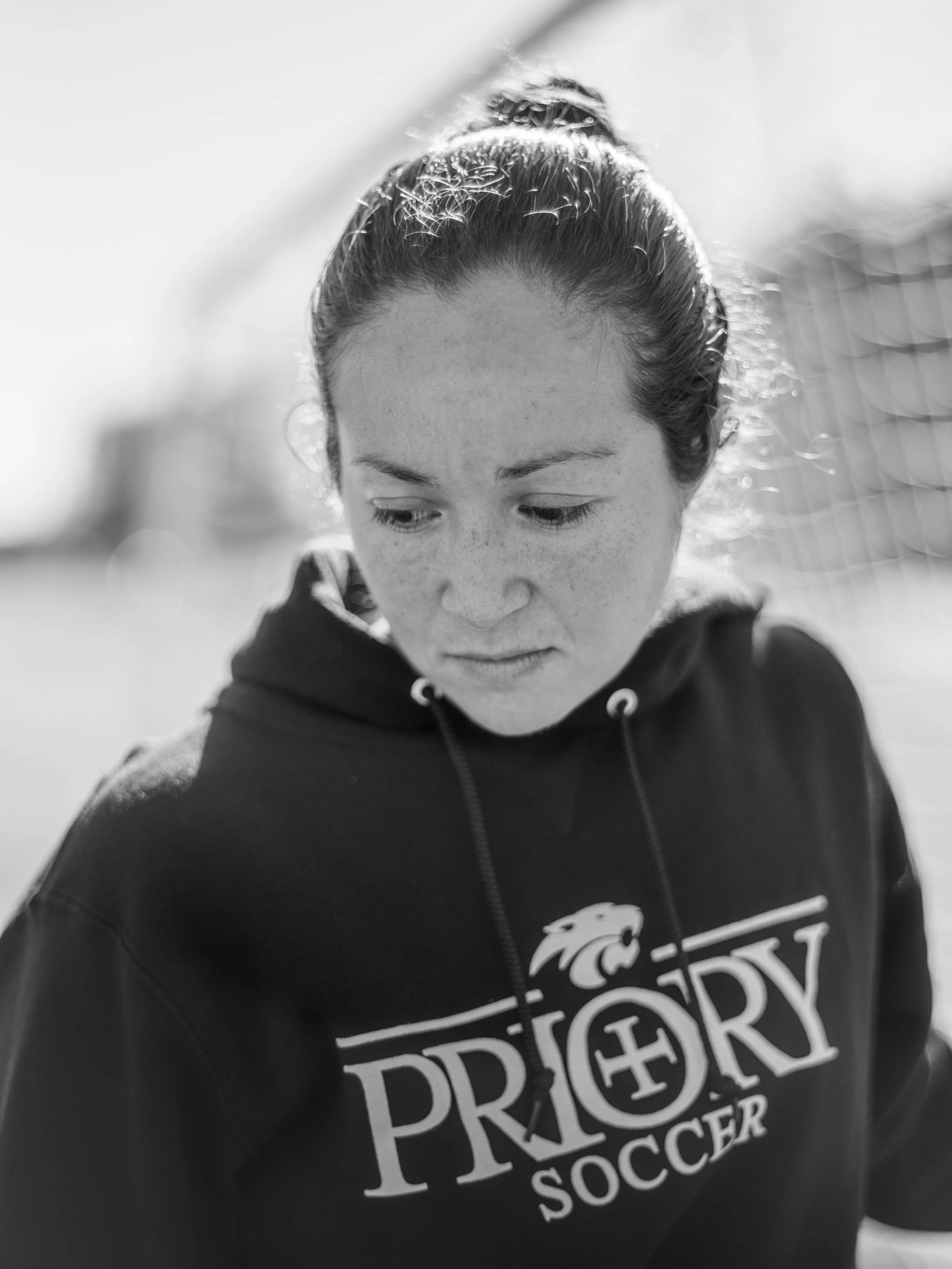 Close-up of a young woman with her hair tied in a bun, wearing a black hoodie with soccer team or sports-related design on the front, outdoors with a blurred background.