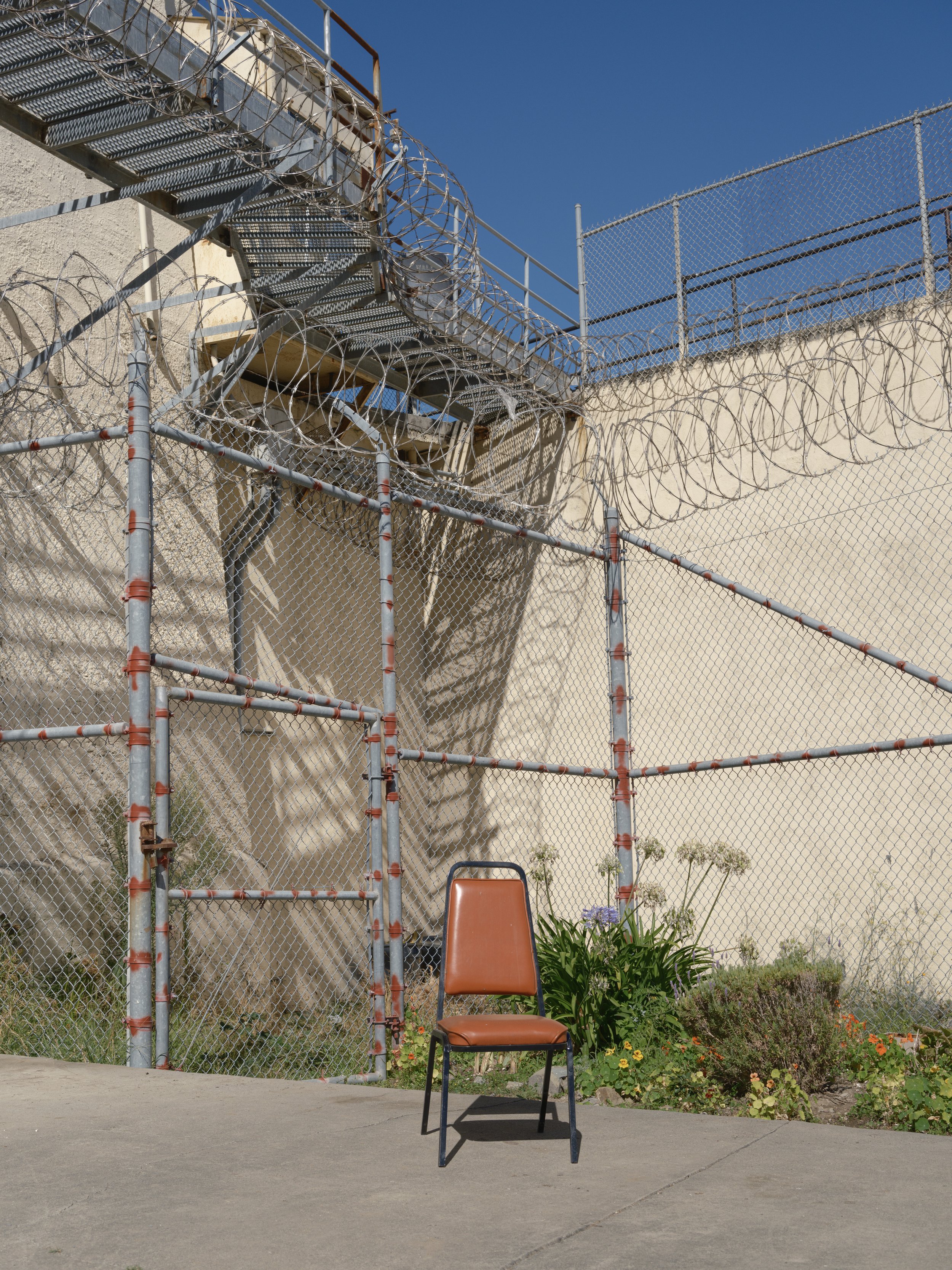 A brown chair with a metal frame sitting on a concrete sidewalk in front of a chain-link fence topped with barbed wire. Behind the fence, there's a beige wall and some green plants with flowers, against a clear blue sky.
