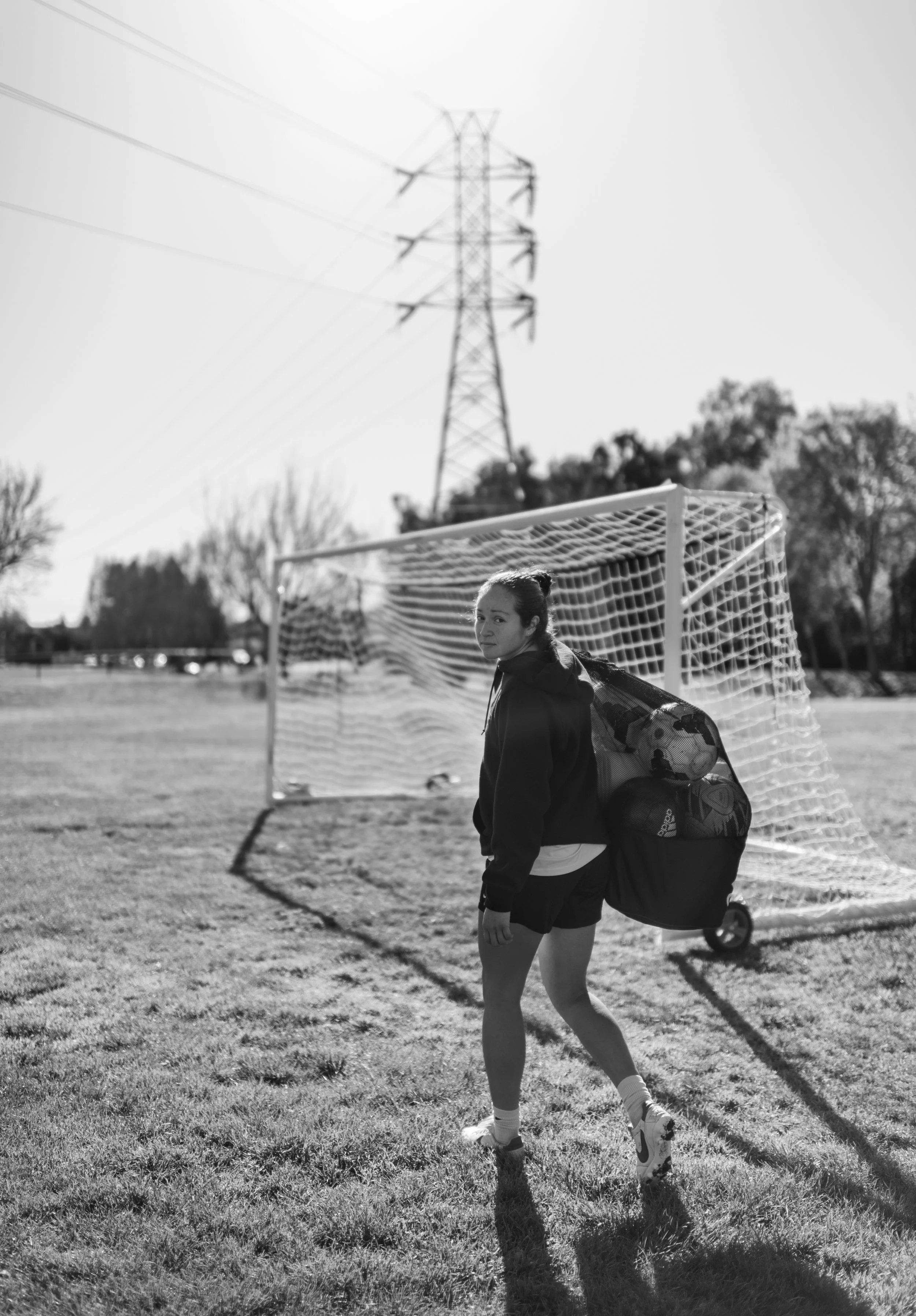 A woman on a soccer field standing near a soccer goal, with a large backpack on her back and a soccer ball in the backpack. She is wearing a jacket and athletic shoes, with trees and a power line tower visible in the background.