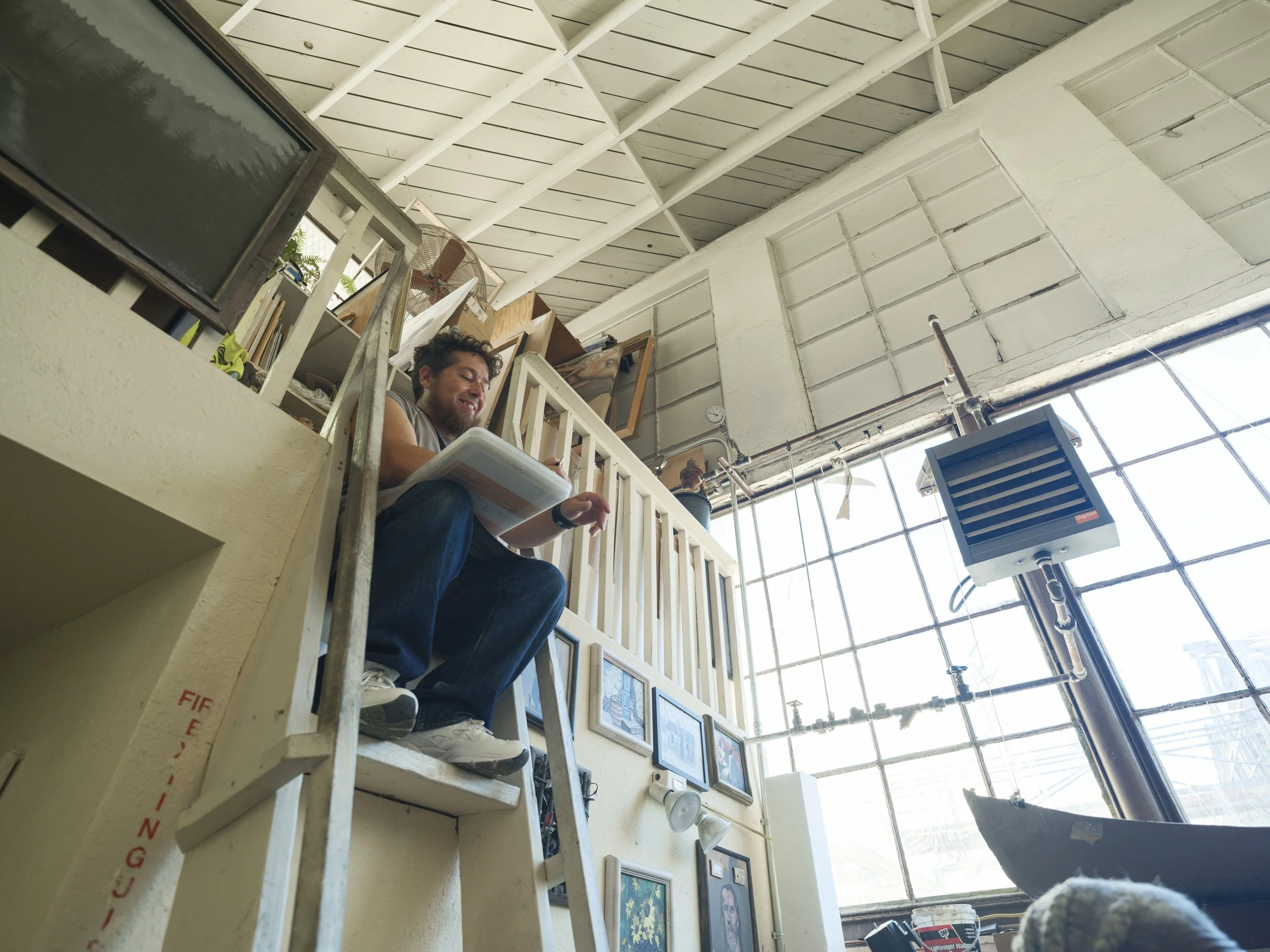 A man with curly hair and a beard sitting on a high platform, smiling and holding a tablet in a bright indoor space with a high ceiling, large windows, and artwork on the walls.