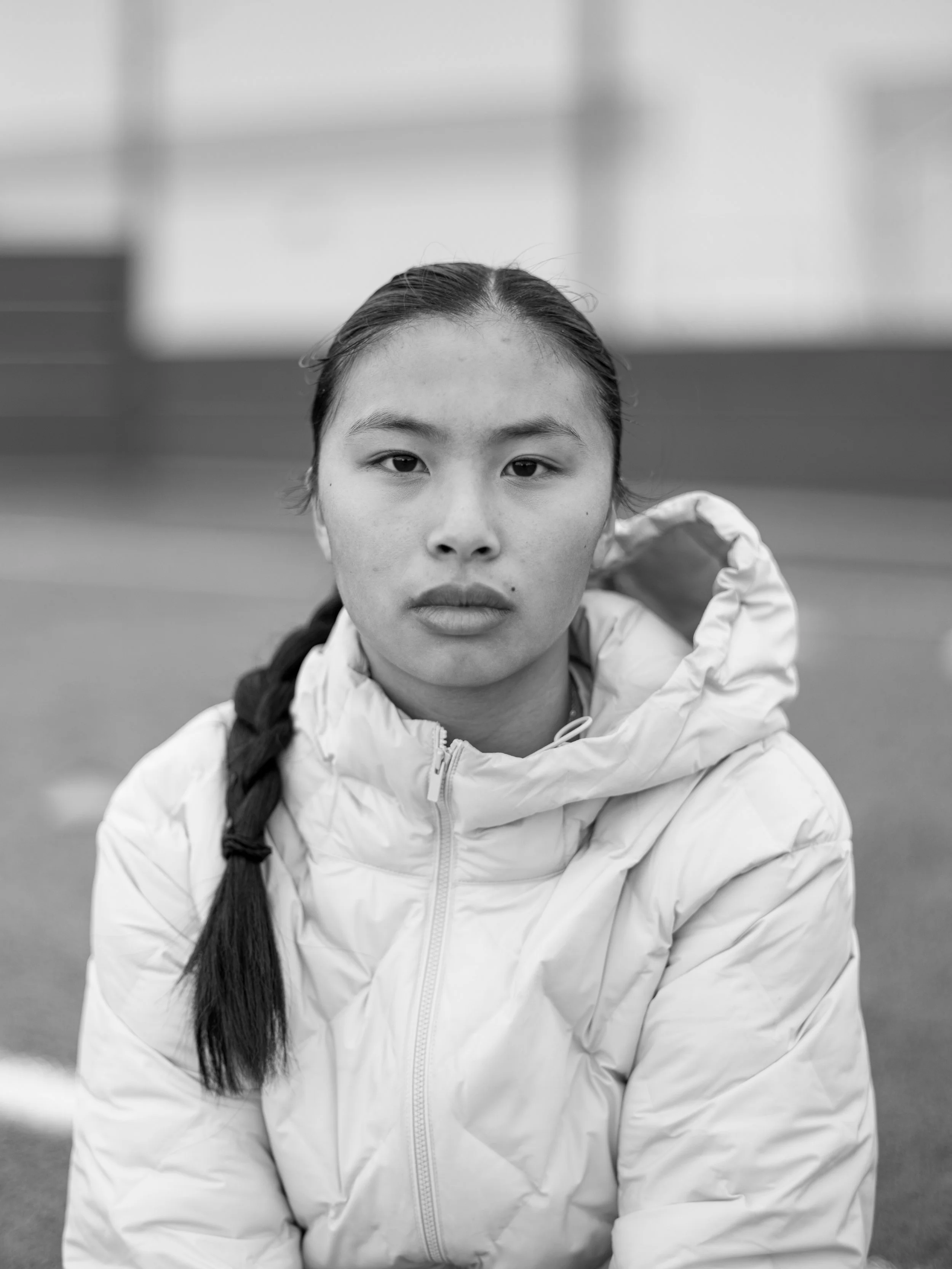 A young woman with long dark hair and a braid, wearing a white puffy jacket, looking directly at the camera in an outdoor setting.