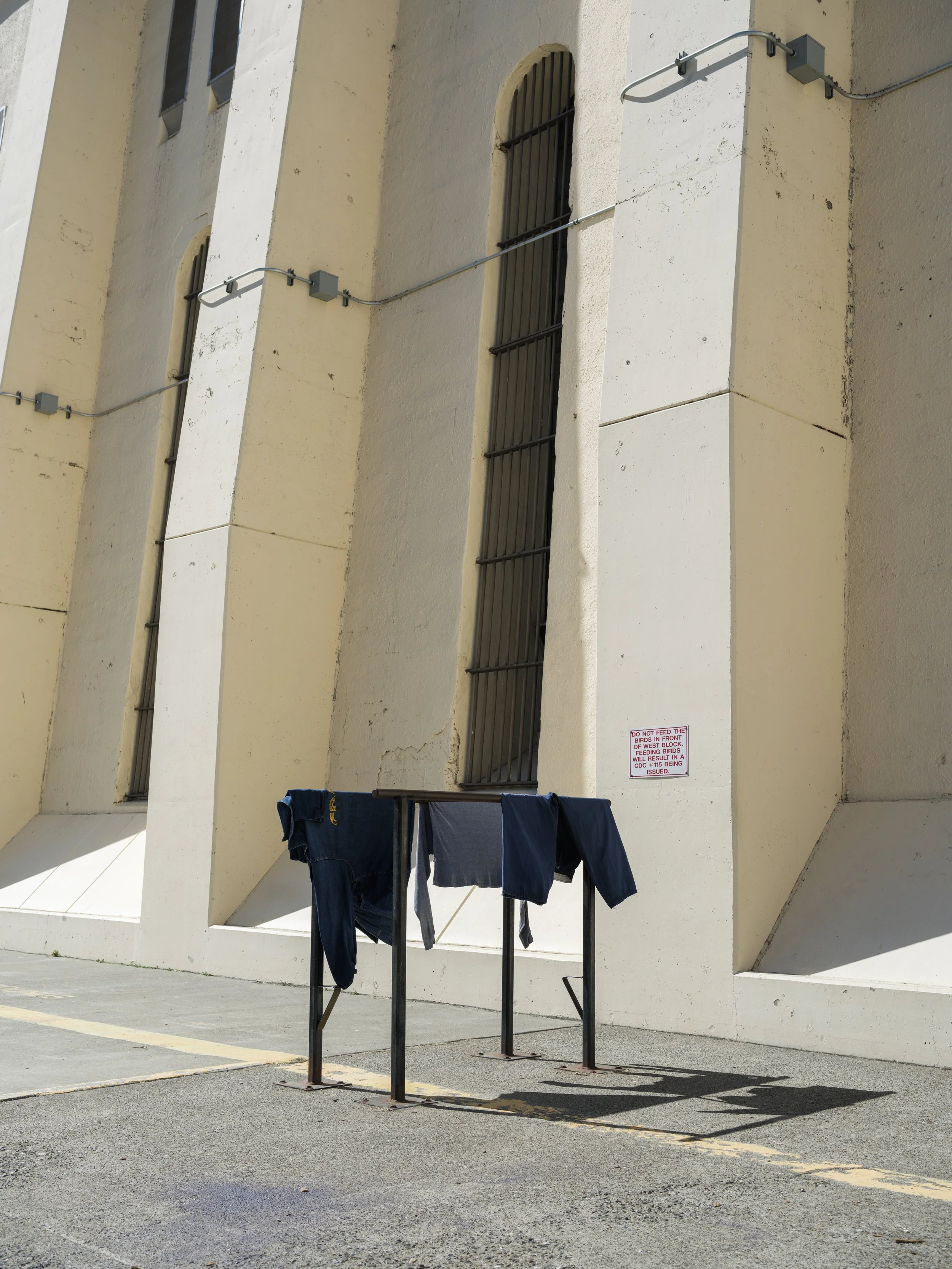 Clothes hanging on a drying rack outside a building with beige walls and small windows. A sign on the wall warns against feeding birds.