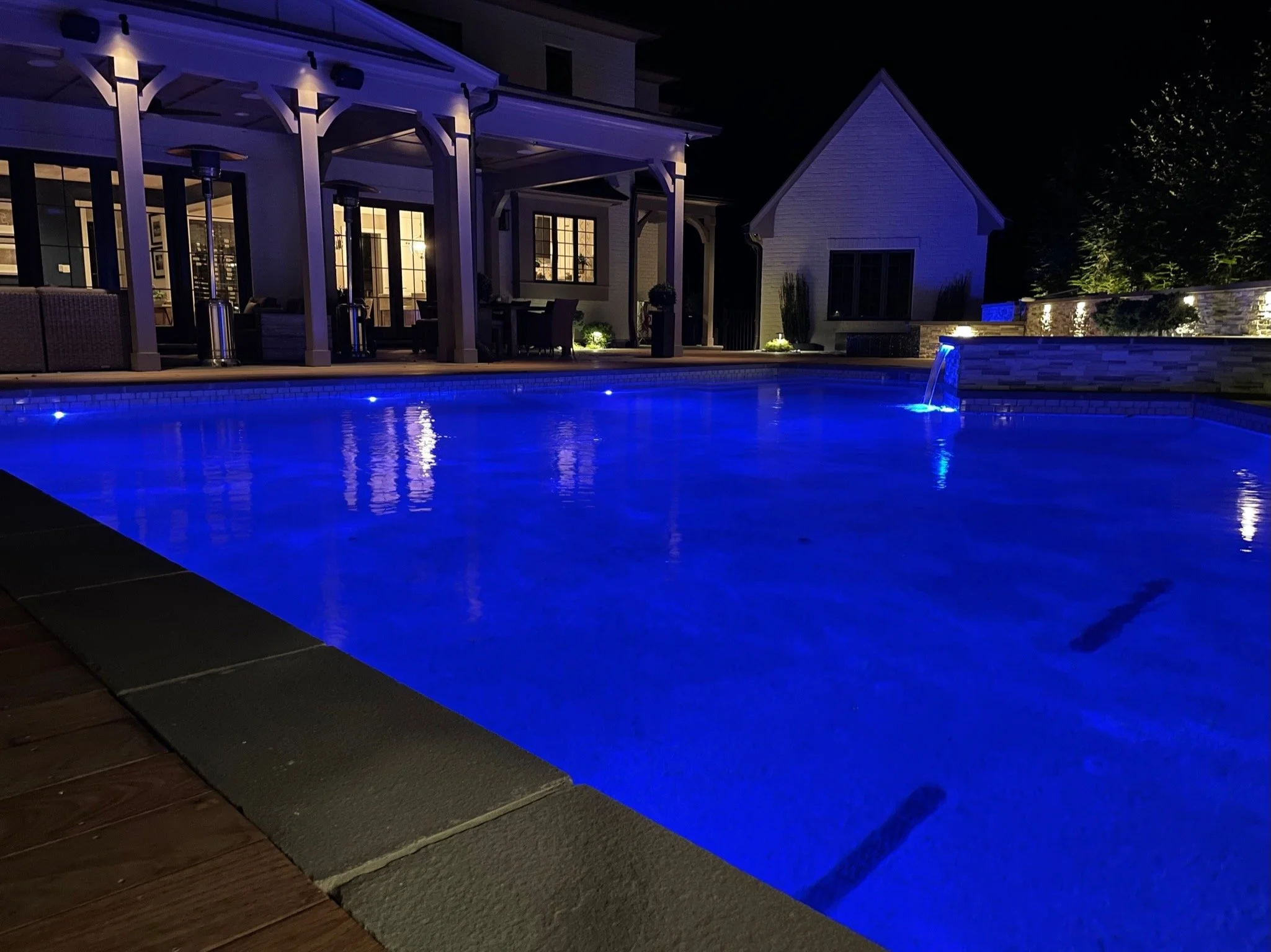 Pool illuminated at night beside a covered patio and stone water feature.