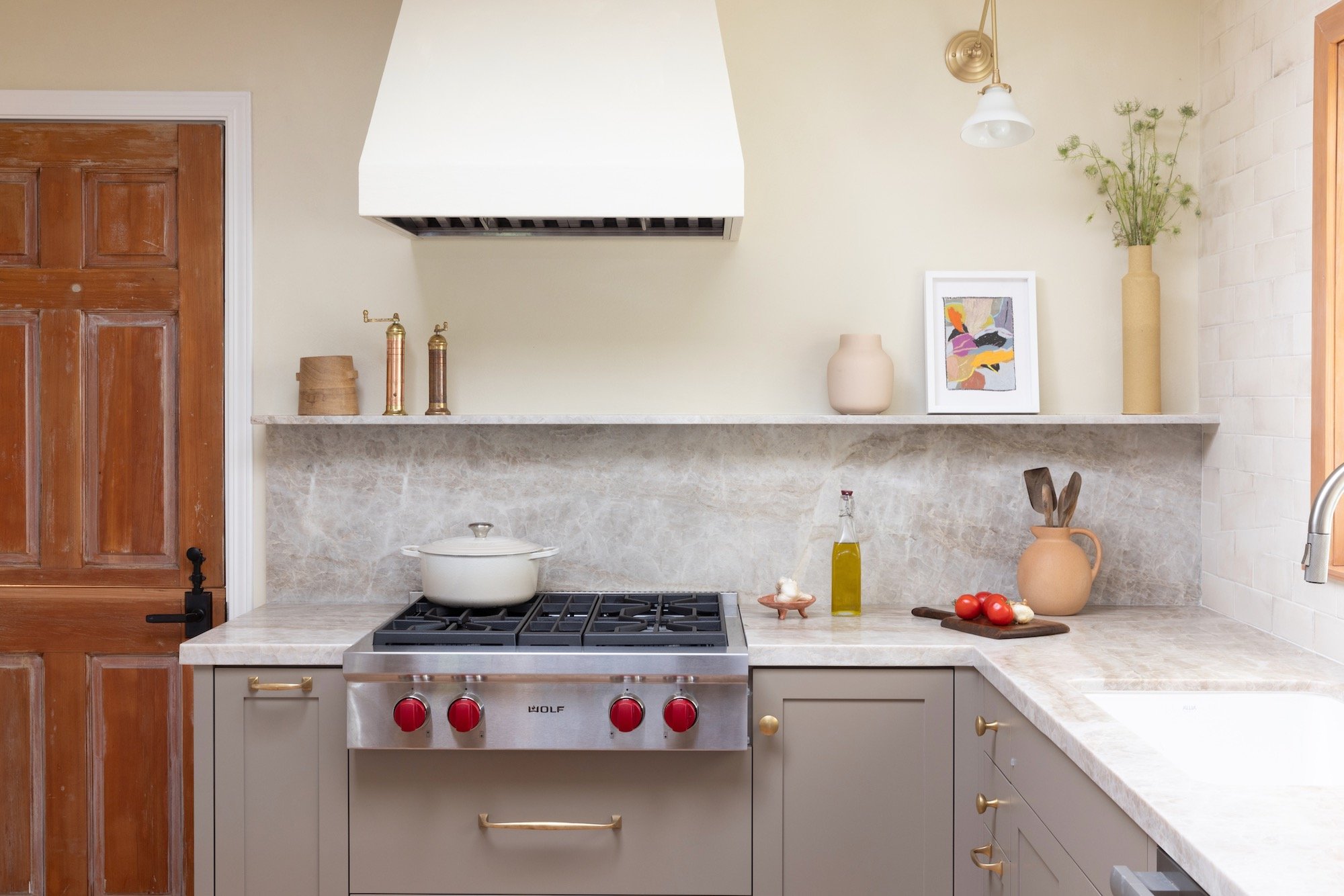 A modern kitchen with beige cabinets, a marble countertop, a stove with a white pot, and decorative items including vases, framed artwork, and fresh tomatoes.