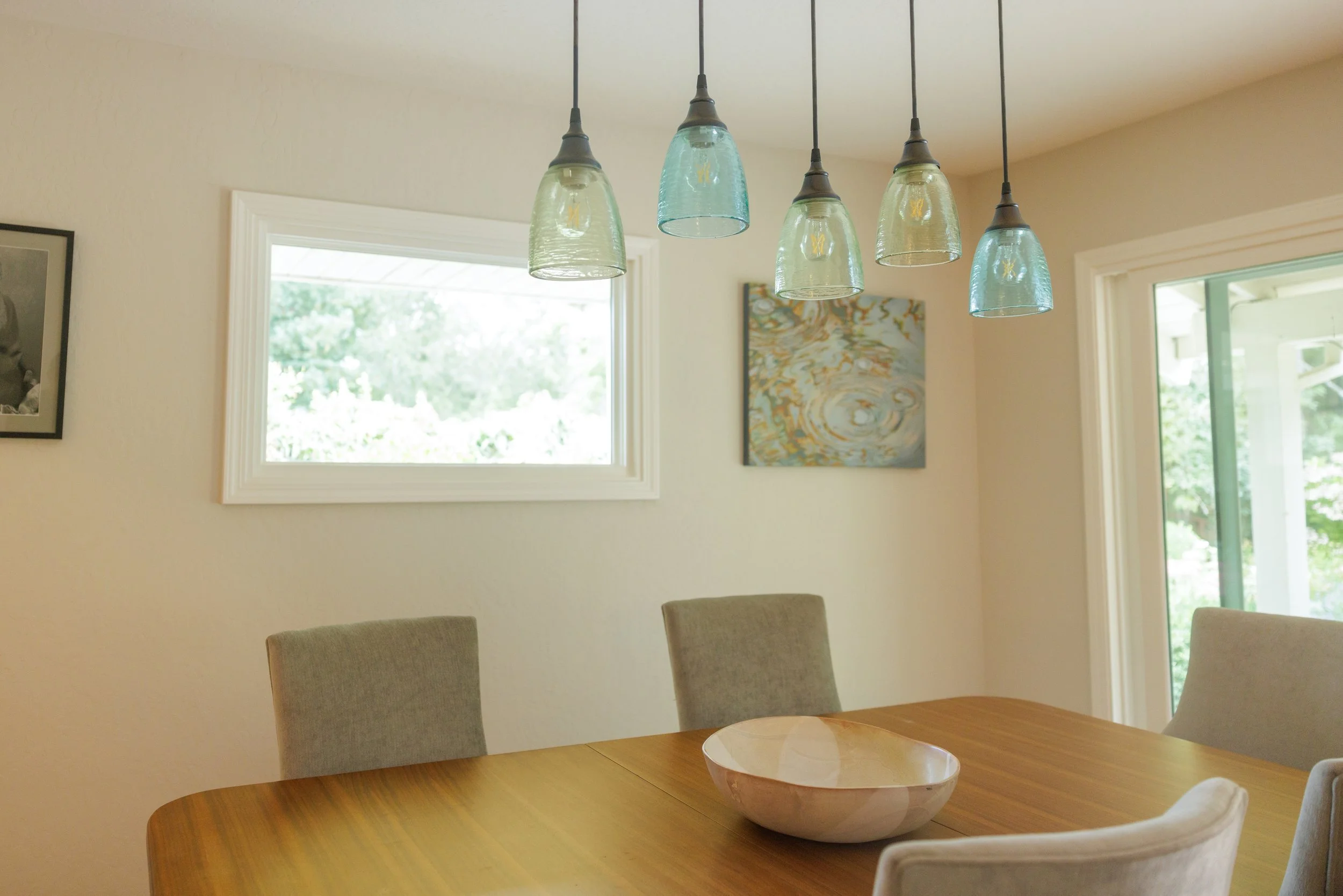 A dining room with a wooden table, beige chairs, a bowl in the center, two windows, wall art, and a five-glass pendant light fixture above the table.