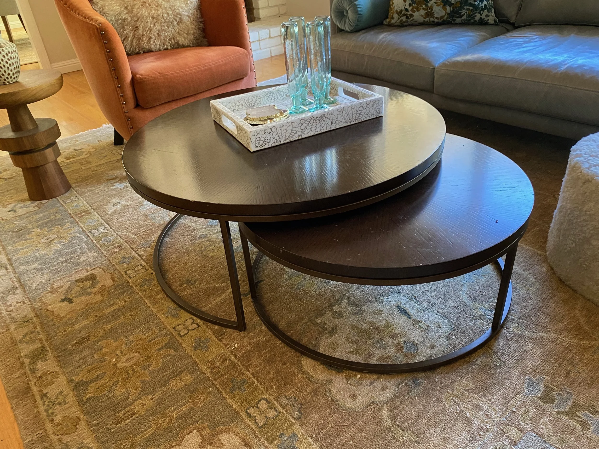 Two nested oval wooden coffee tables with black legs, one placed on top of the other. On the top table, there is a white decorative tray with three tall blue vases and a couple of small decorative items. The tables are in a living room with a patterned area rug, a burnt orange armchair, a fireplace, a gray sofa, a fluffy white ottoman, and a wooden side table with a decorative vase.
