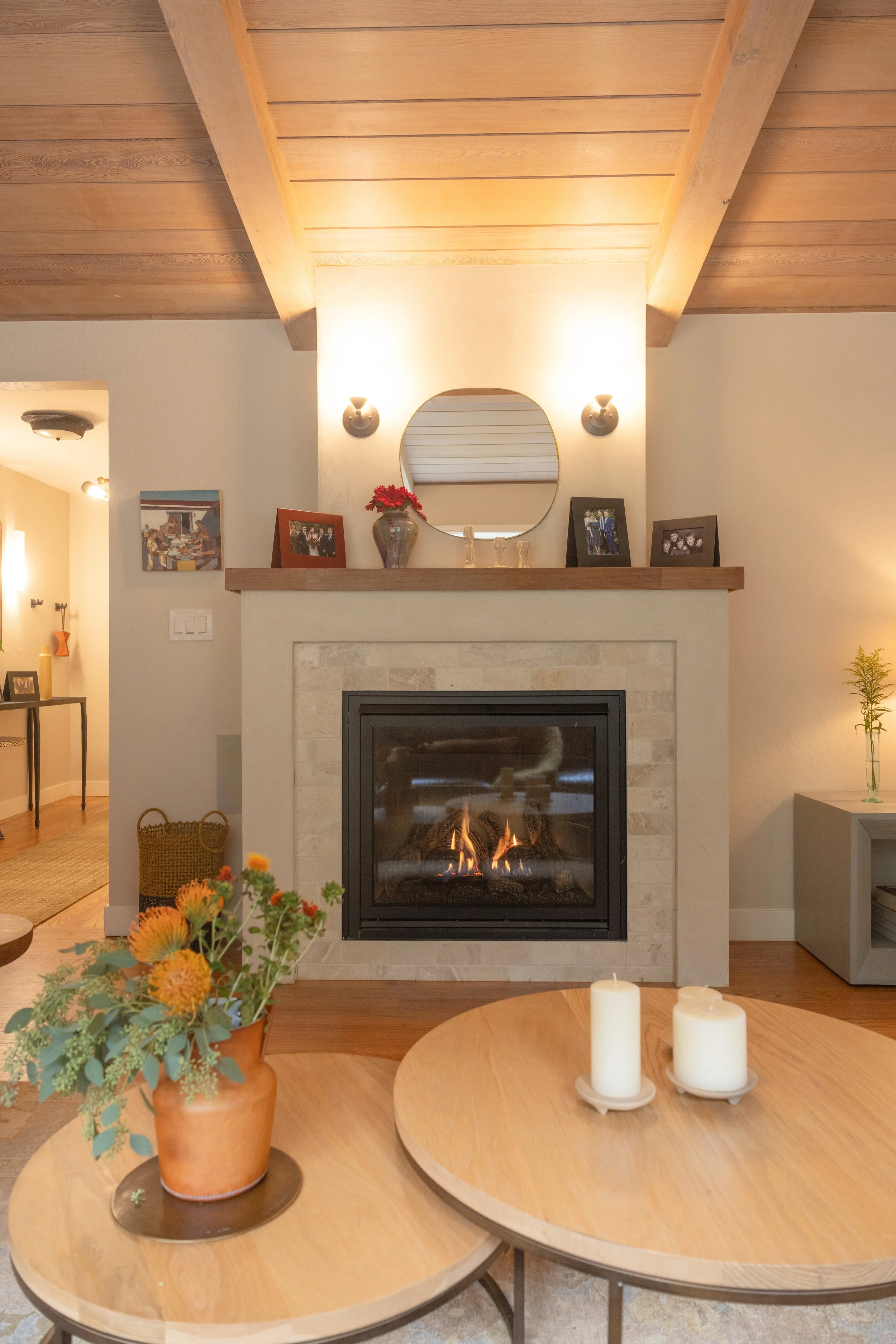 Living room with a fireplace, decorated with framed photos, a mirror, and a vase with red flowers on the mantel. In front, two round wooden tables hold a vase with flowers and three candles. The room has a wooden ceiling and a side table with a plant.