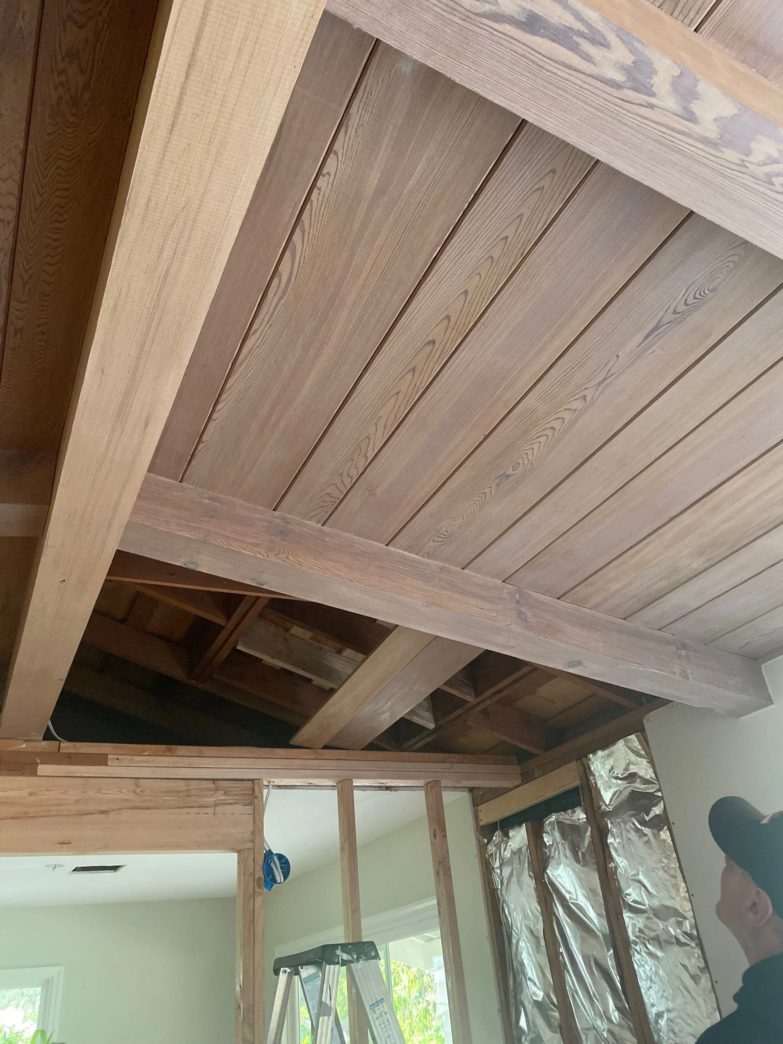 Interior of a house under construction, showing exposed wooden ceiling beams and partially framed walls, with a person wearing a cap in the lower right corner.
